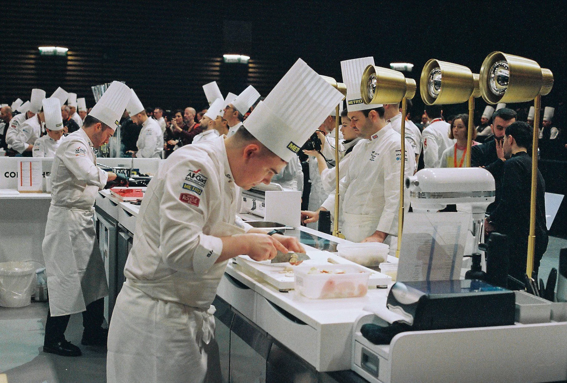 A group of professional chefs preparing food at a culinary competition or event, wearing white uniforms and tall chef hats, working at a stainless steel counter in a large, well-lit space.