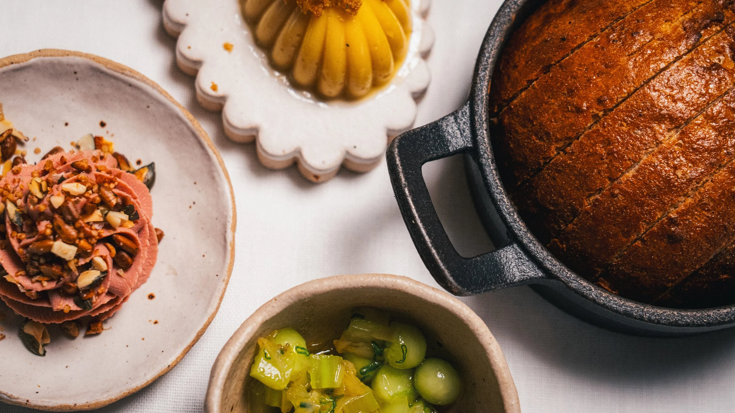 A variety of dishes including sliced meat with chopped nuts, pickled vegetables, a loaf of baked bread, and condiments on white plates and bowls.