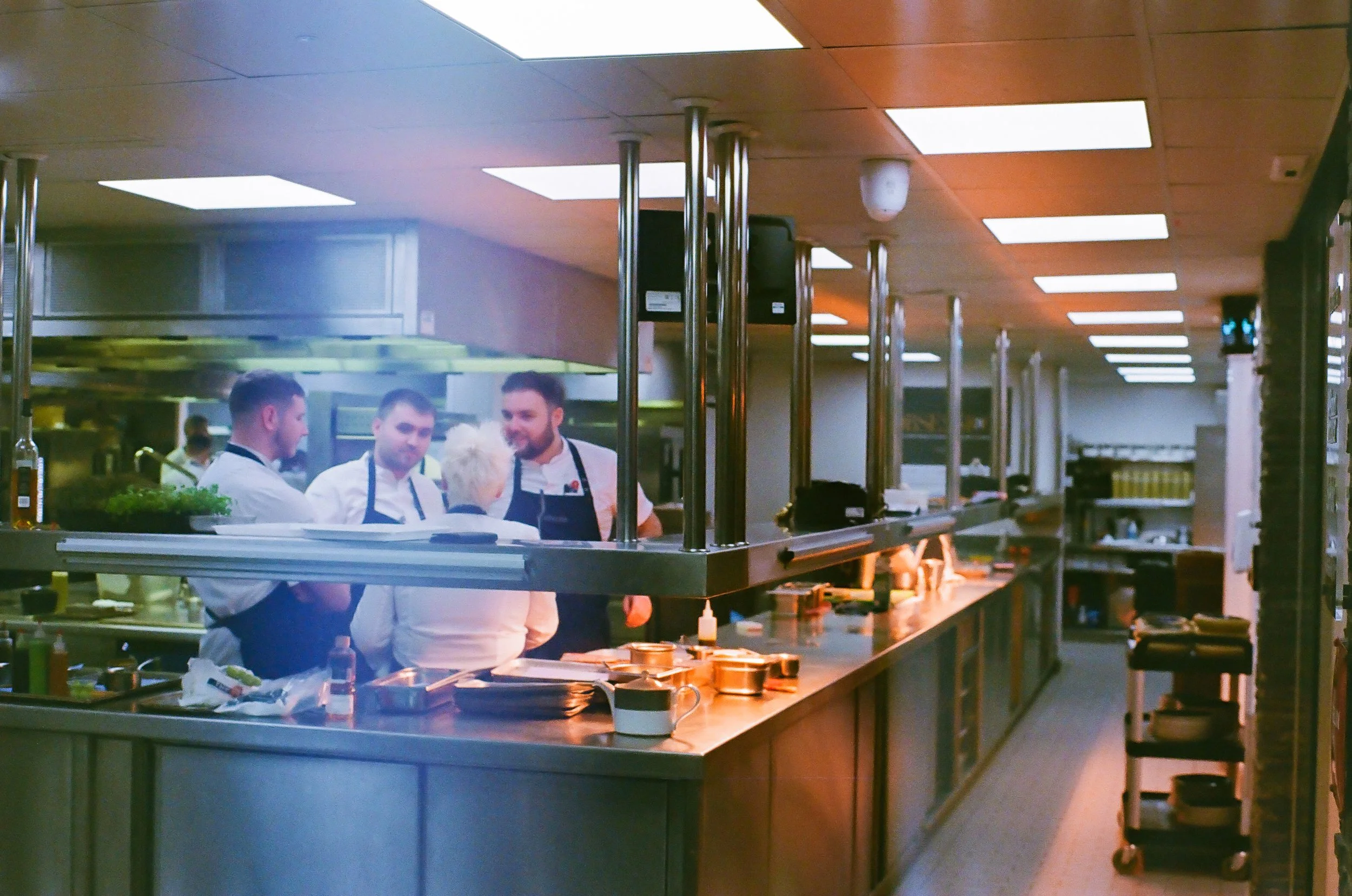 Kitchen scene with three chefs in white uniforms and aprons working behind a stainless steel counter, with overhead lighting and kitchen equipment.