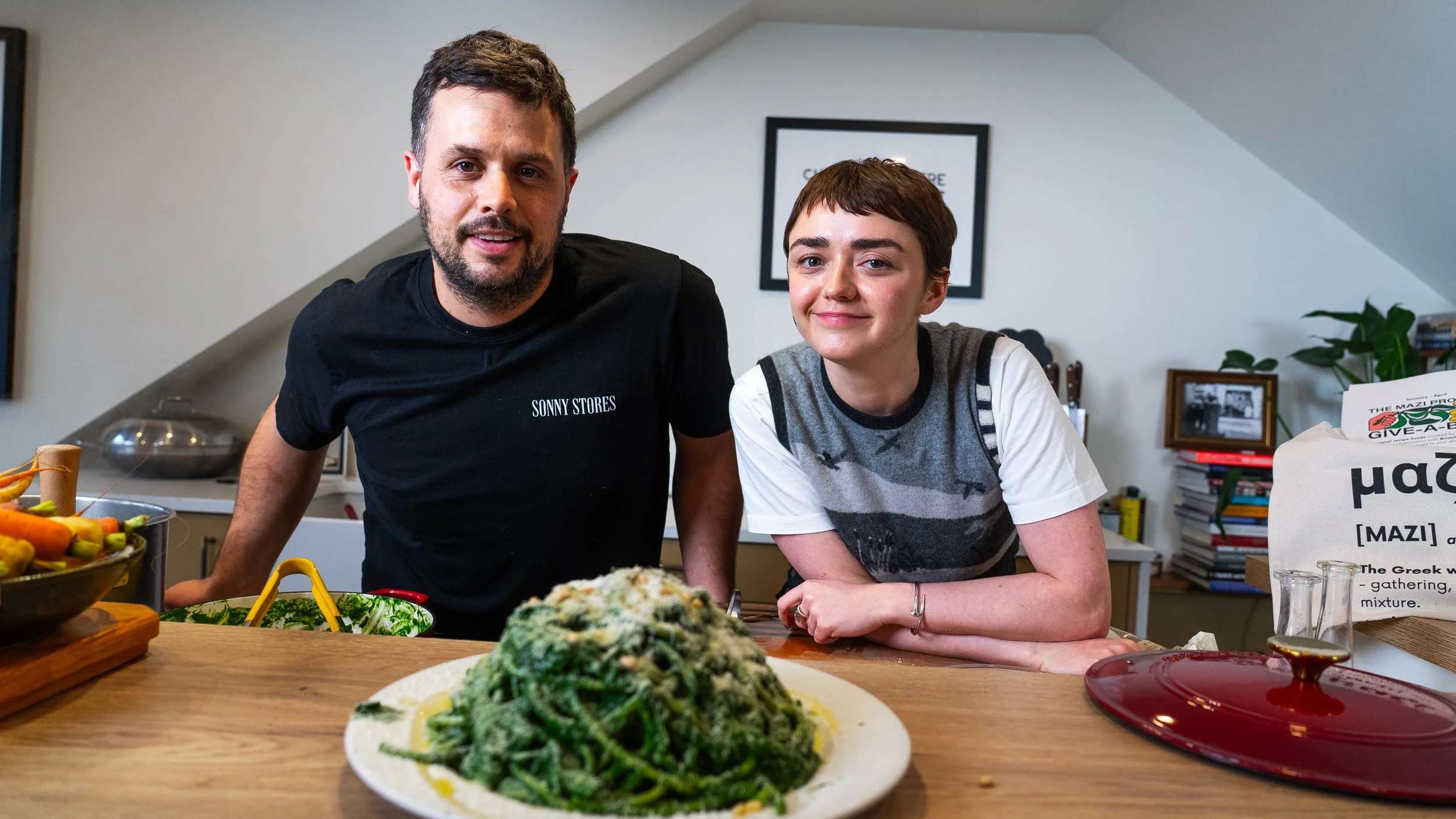 A man and a woman with short hair are leaning on a kitchen countertop with food around them, smiling at the camera. In the foreground, there is a plate of green pasta or vegetable dish, and on the left side, a bowl of vegetables. The kitchen has a white wall with picture frames, books, and a plant in the background.