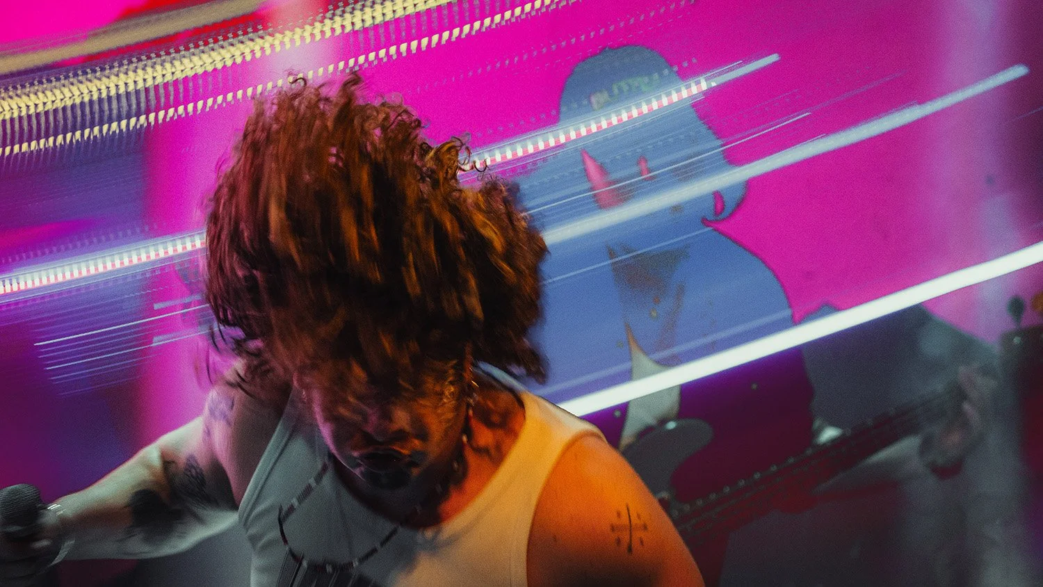 A person with curly red hair singing into a microphone in front of a pink and purple background with streaks of light and digital effects.