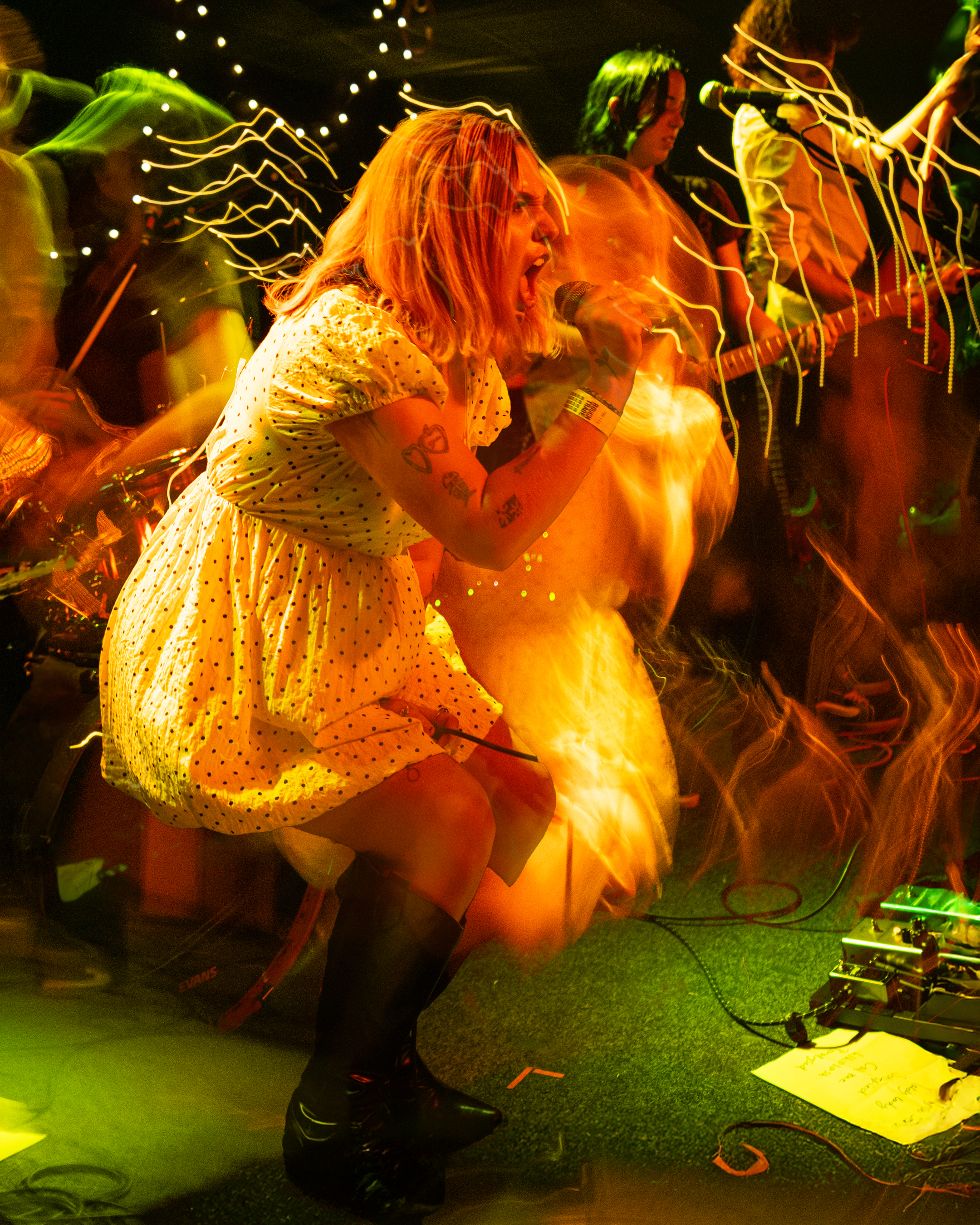 A woman with blonde hair wearing a yellow polka dot dress sings into a microphone on stage, with other band members playing instruments behind her. The scene is vibrant with colorful light streaks and blurred motion, suggesting energetic performance.
