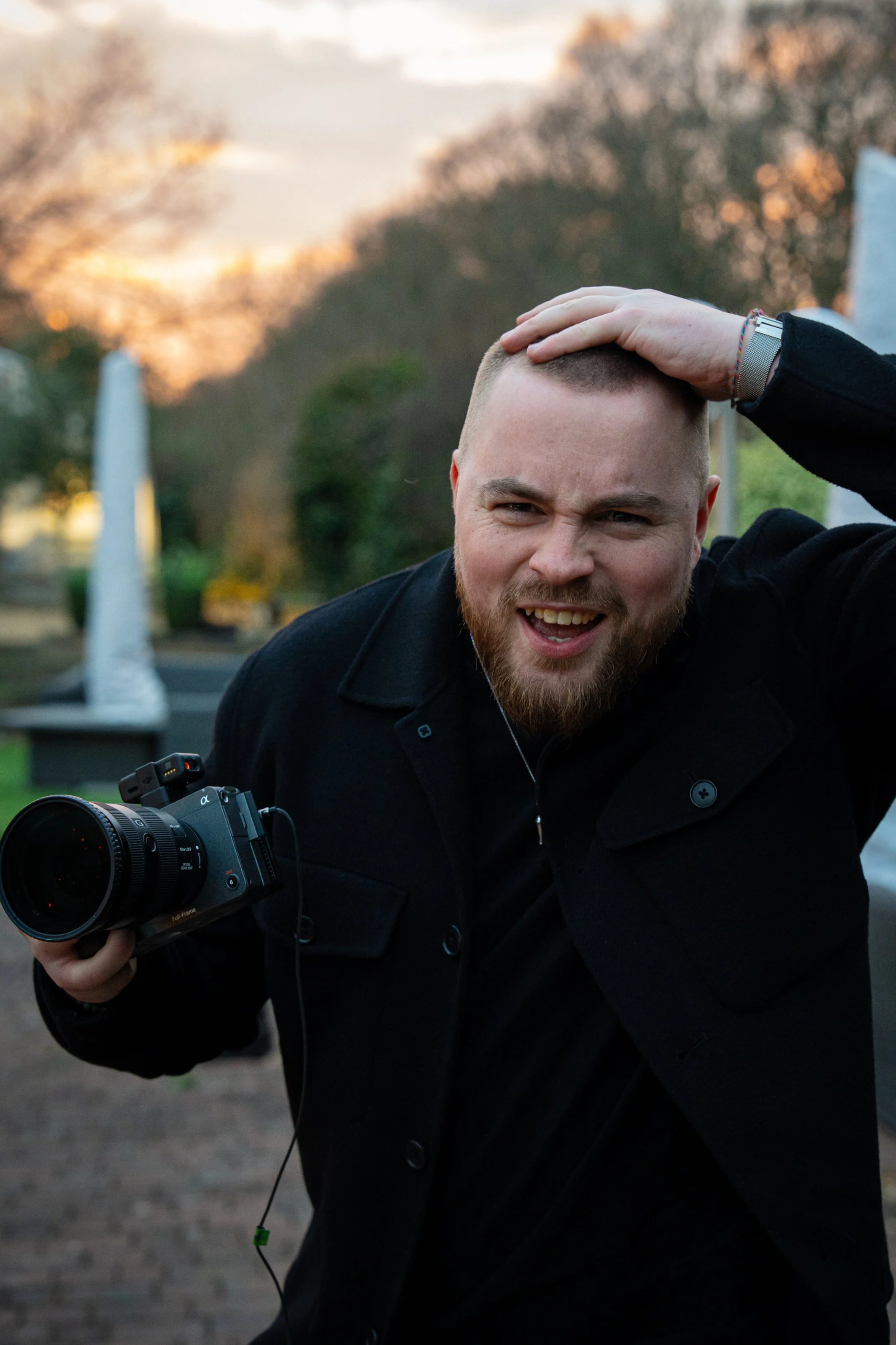 A man with a beard wearing a black coat is holding a camera, with his left hand on his head, smiling in an outdoor setting during sunset with trees in the background.