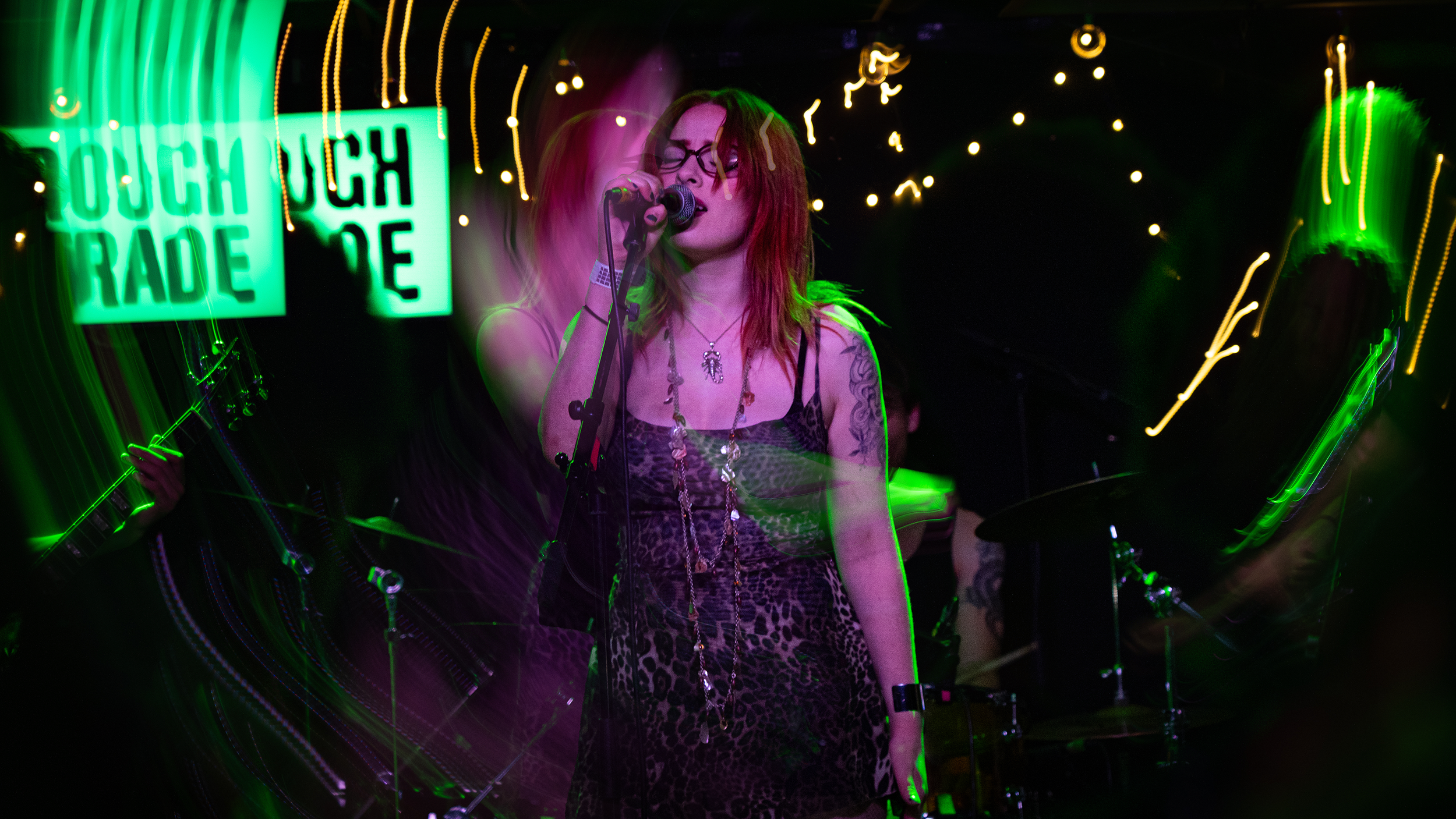 A female singer with red hair, glasses, and tattoos singing into a microphone on stage with neon green signs and colorful lights.