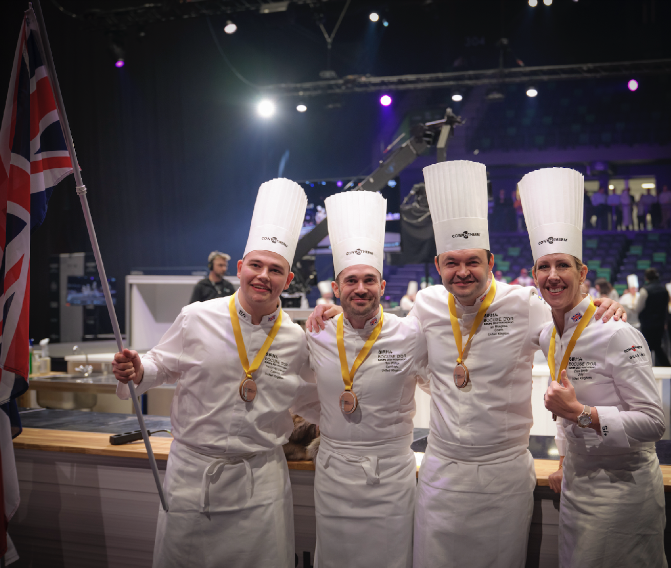 Four chefs in white uniforms and tall chef hats, each wearing a medal, standing together and smiling with arms around each other. One chef on the far left is holding a Union Jack flag. The background suggests a professional kitchen or culinary competition setting with professional equipment and a camera crane.
