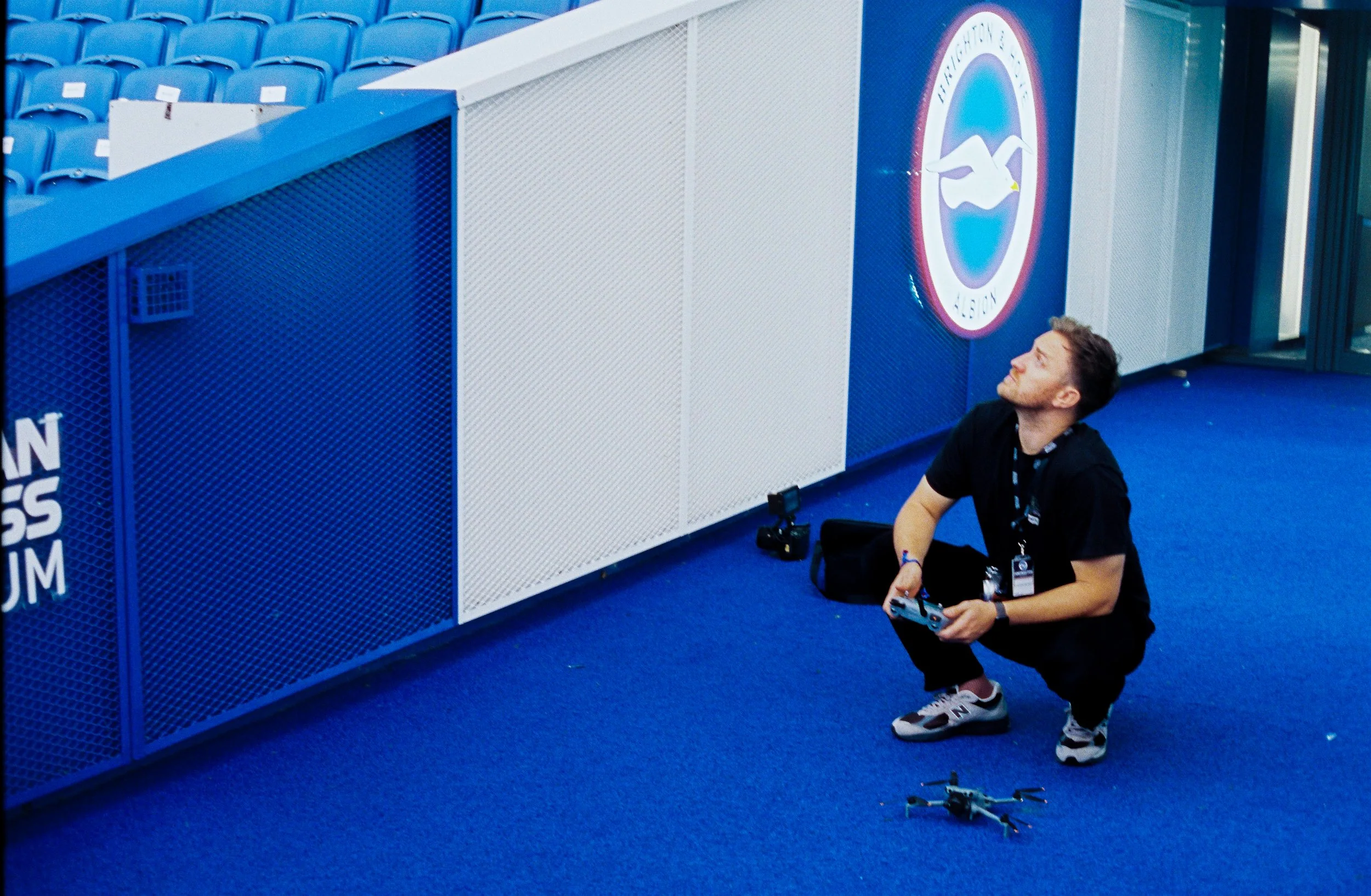 A man crouching on a blue sports arena floor, holding a remote control drone, with blue stadium seats and a large team logo sign in the background.