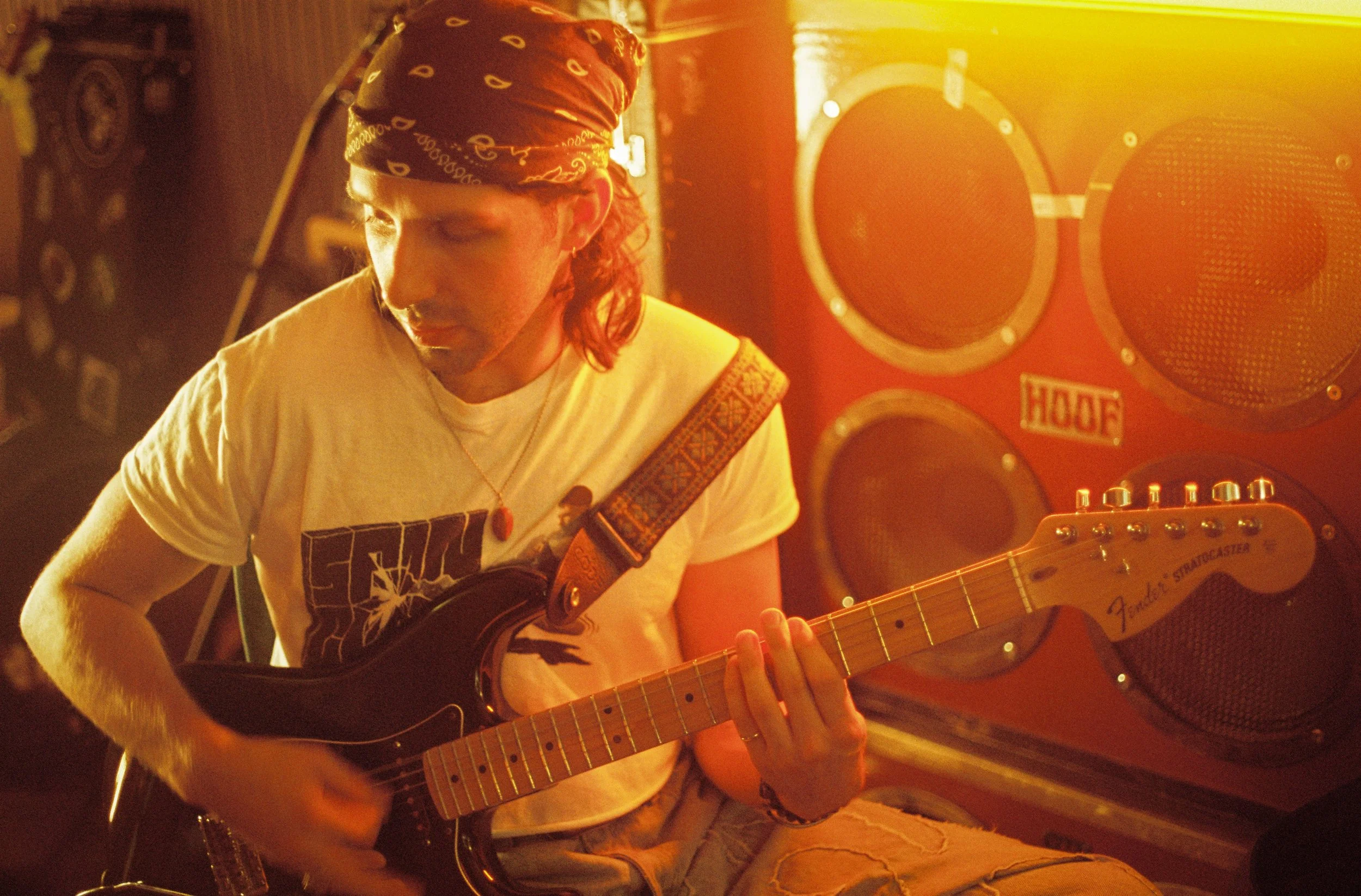 A person with long curly hair wearing a bandana, playing an electric guitar in a warmly lit room with large speakers in the background.