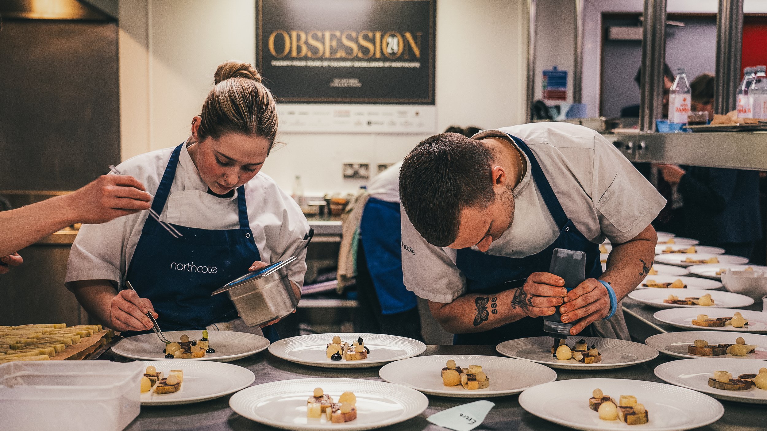Two chefs in a professional kitchen carefully inspect meticulously plated desserts on a counter, with numerous finished dessert plates arranged in rows in front of them.