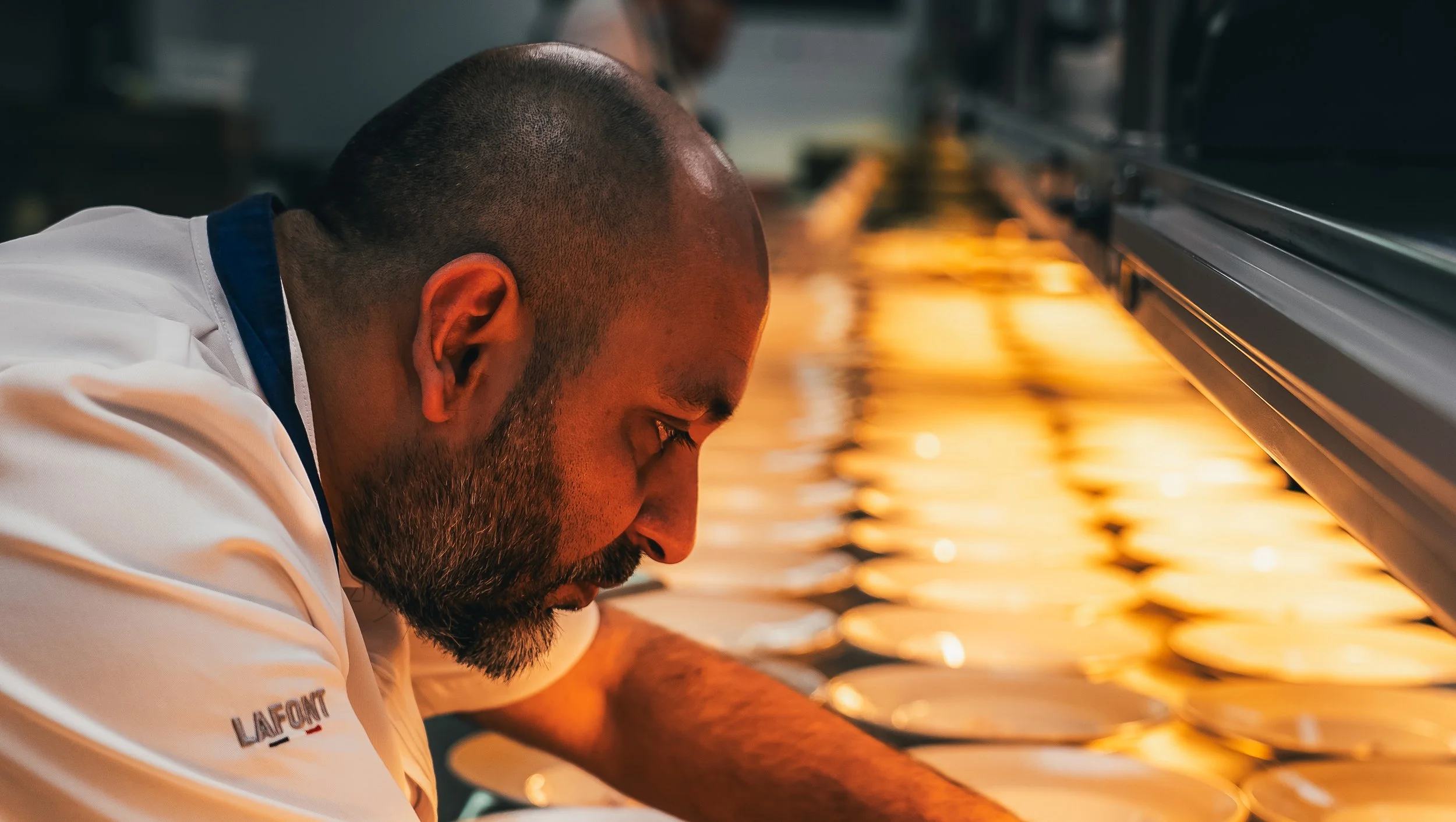A man with a beard and a shaved head leaning over a counter with a warm, glowing light reflected on its surface.