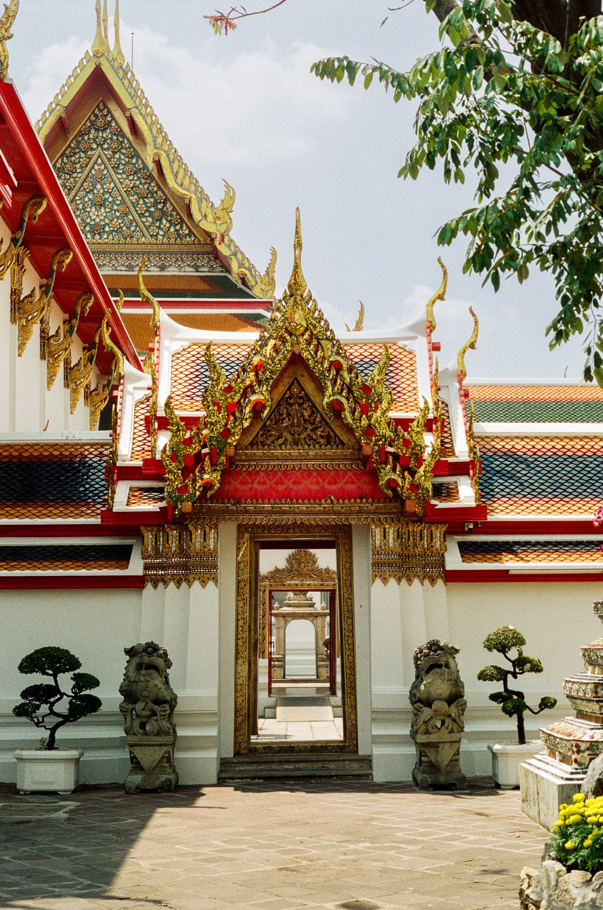 Traditional Thai temple with ornate, golden detailing, red and green tiled roof, stone lion statues, and manicured plants in pots, under a cloudy sky.