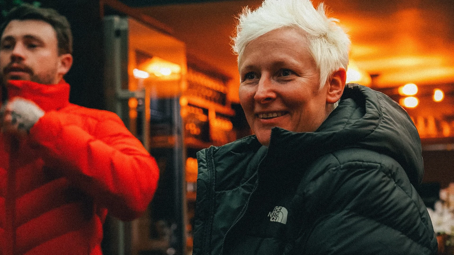 Two smiling individuals, one with platinum blonde hair and a black North Face jacket, and the other with facial hair and wearing a red puffer jacket, engaged in conversation indoors with warm lighting in the background.