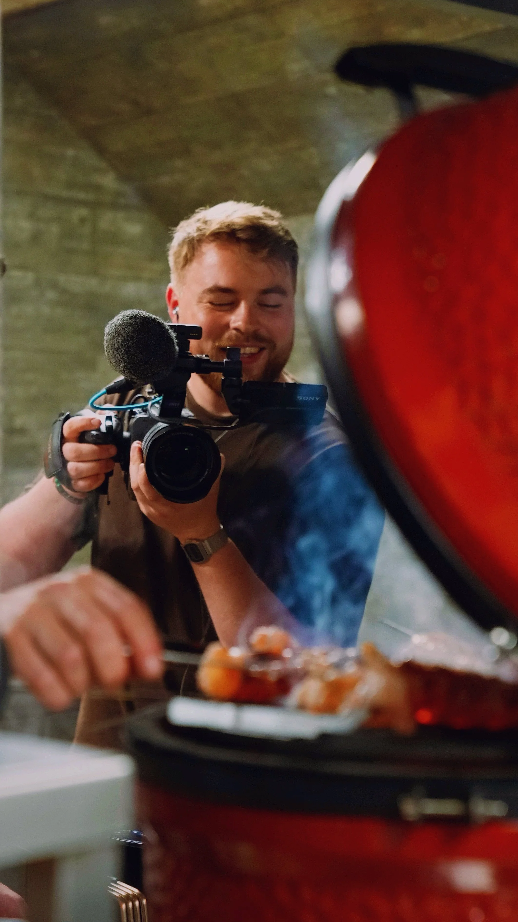 A man smiling and taking a photo with a camera in a kitchen setting, with a food dish in the foreground.