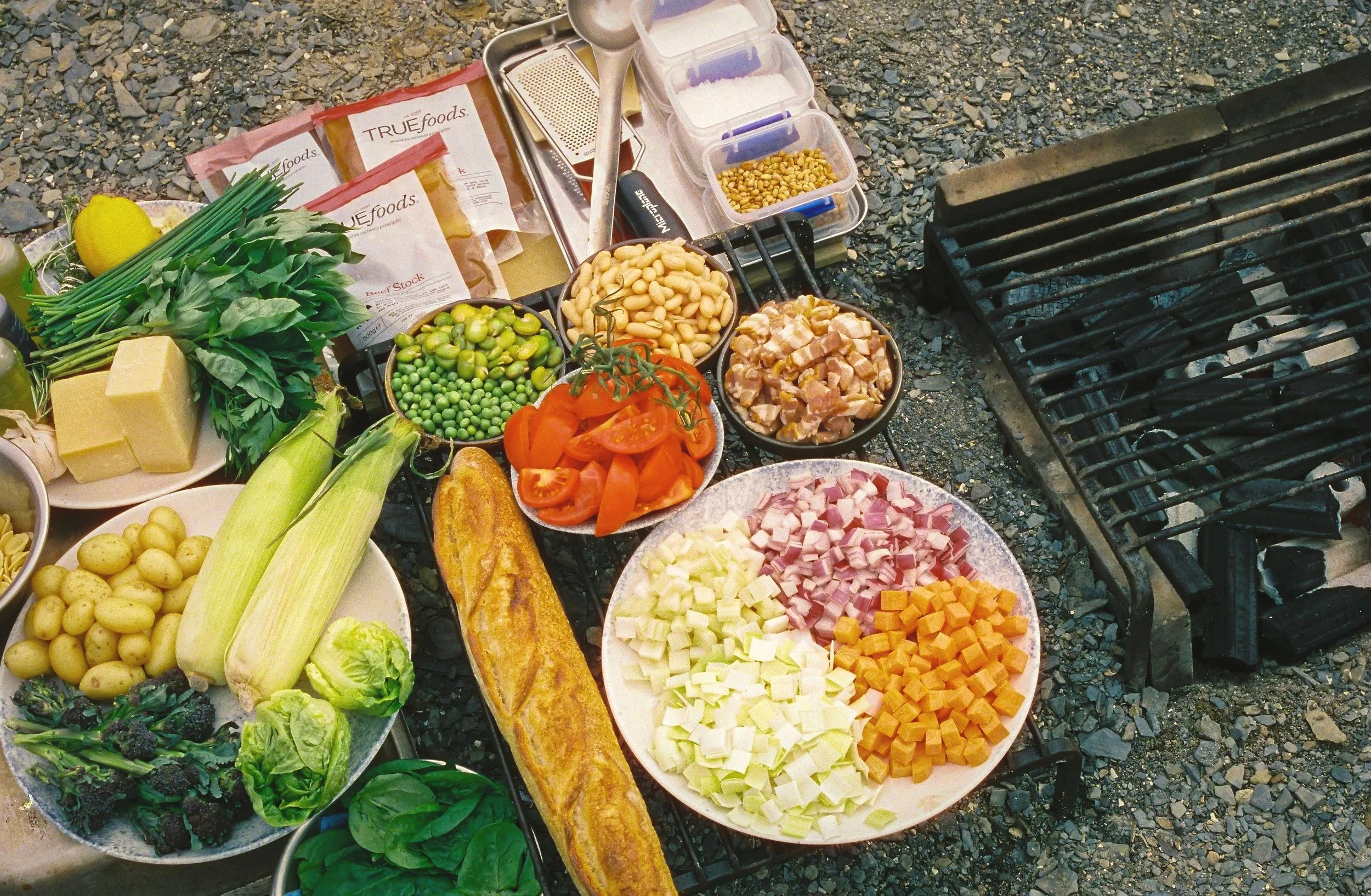 Various fresh vegetables and ingredients laid out on a grill and nearby plates, including cherry tomatoes, cubed cheese, diced ham, potatoes, green beans, lettuce, and herbs, with a grilling grate to the right.