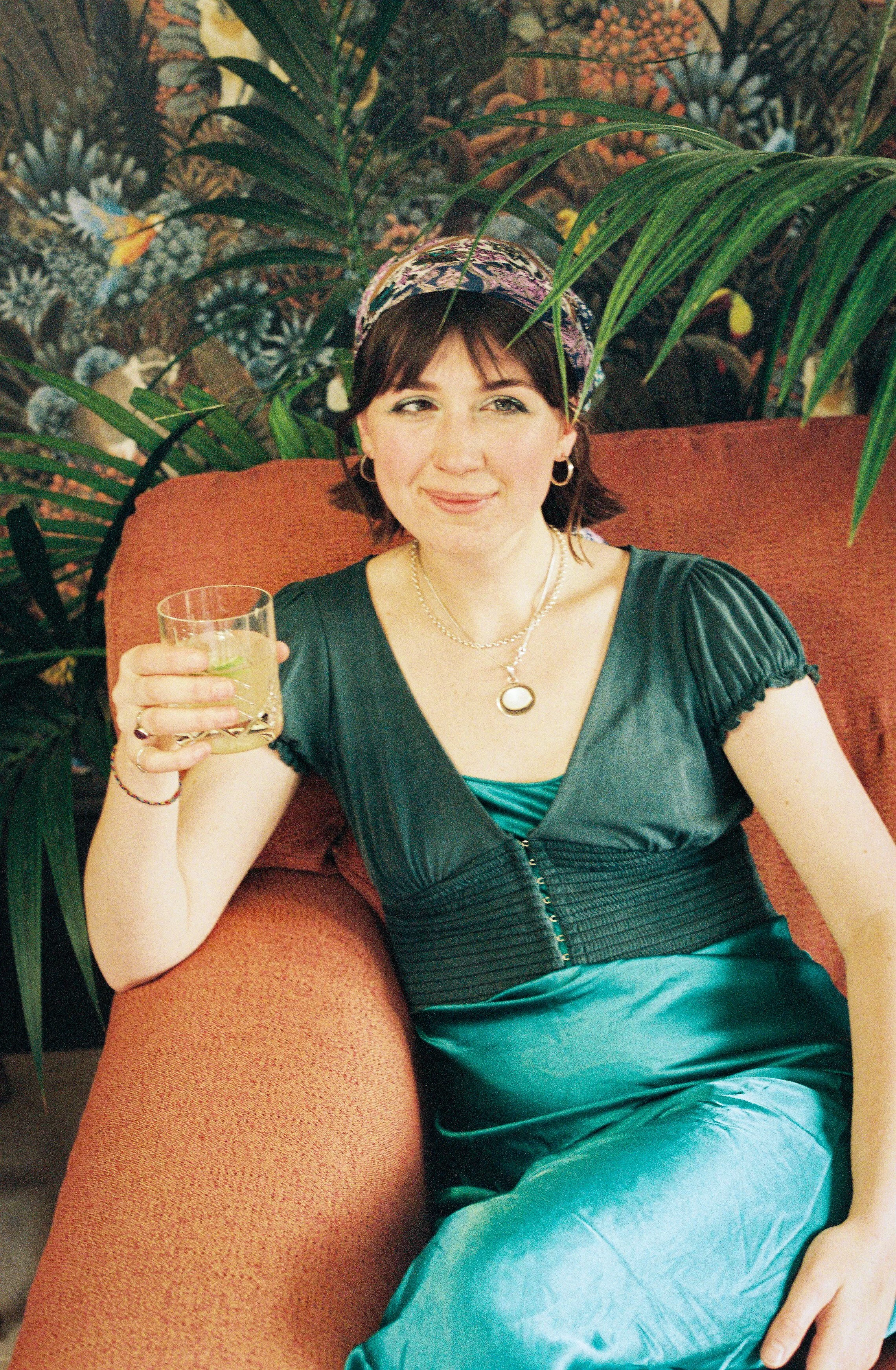 A woman sitting on an orange couch, wearing a dark green dress with a zipper, a patterned headscarf, and jewelry, holding a glass of drink, surrounded by green plants in the background.