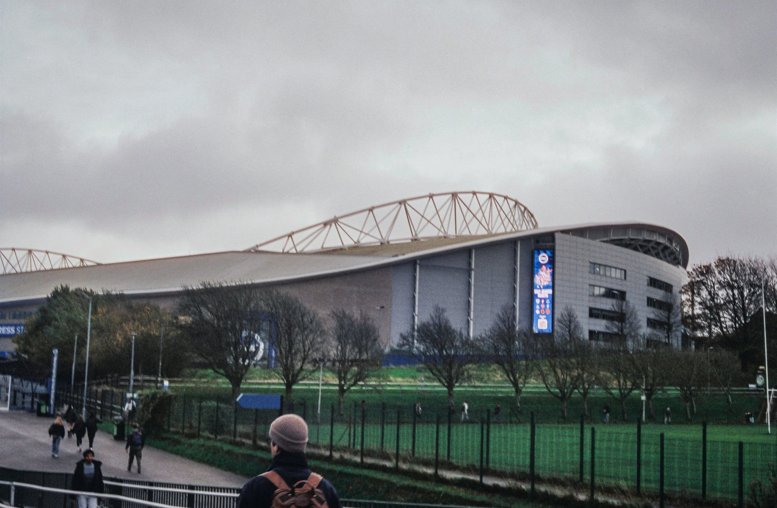 The image shows a stadium or arena with a modern design, featuring a curved roof and lattice structure. There are overcast skies above, and trees and people walking in the foreground.