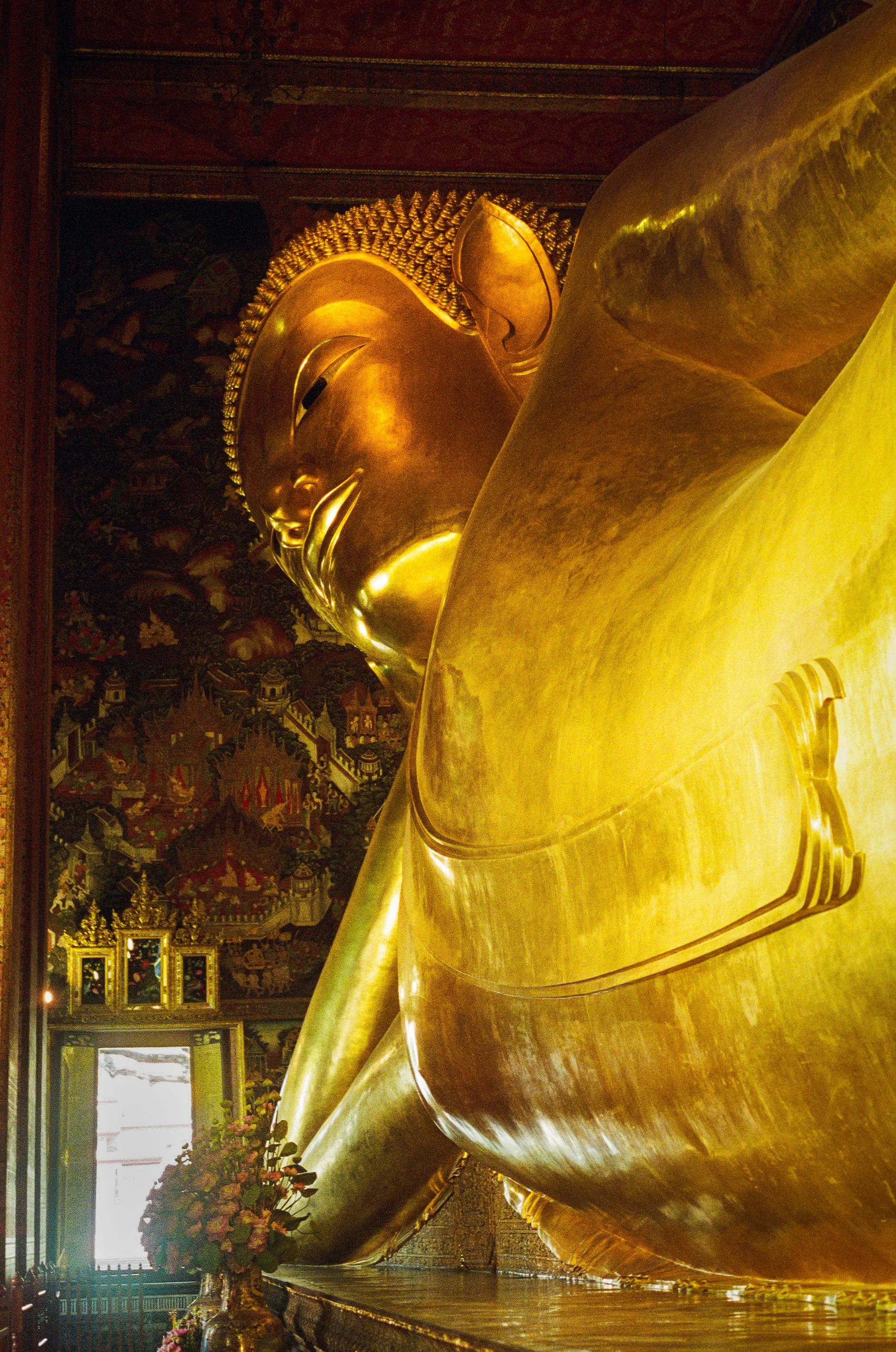 A large reclining golden Buddha statue inside a decorated temple, with ornate paintings and decorations on the walls.