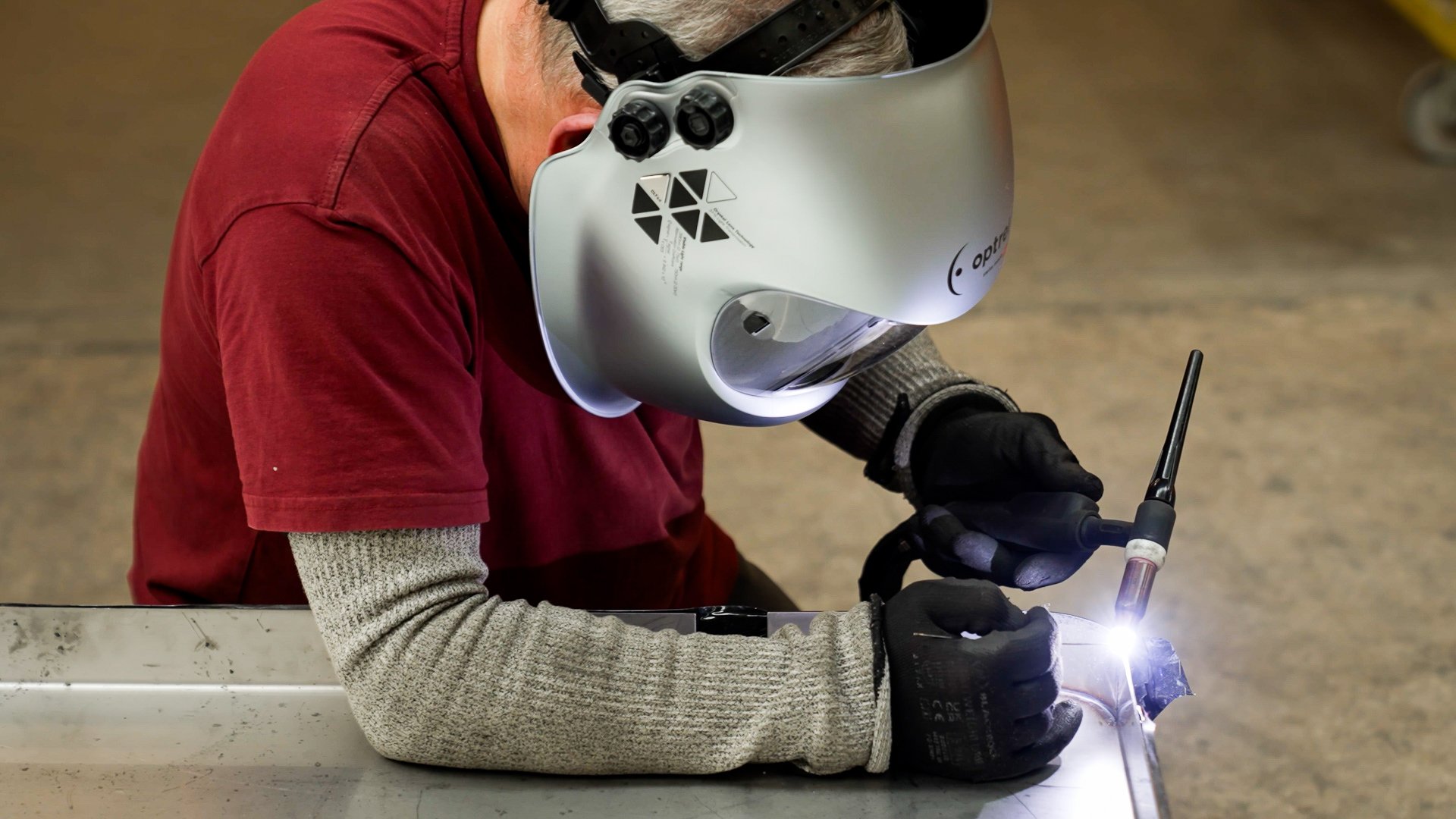 Person wearing a welding helmet and gloves practicing welding on a metal surface in a workshop or industrial setting.