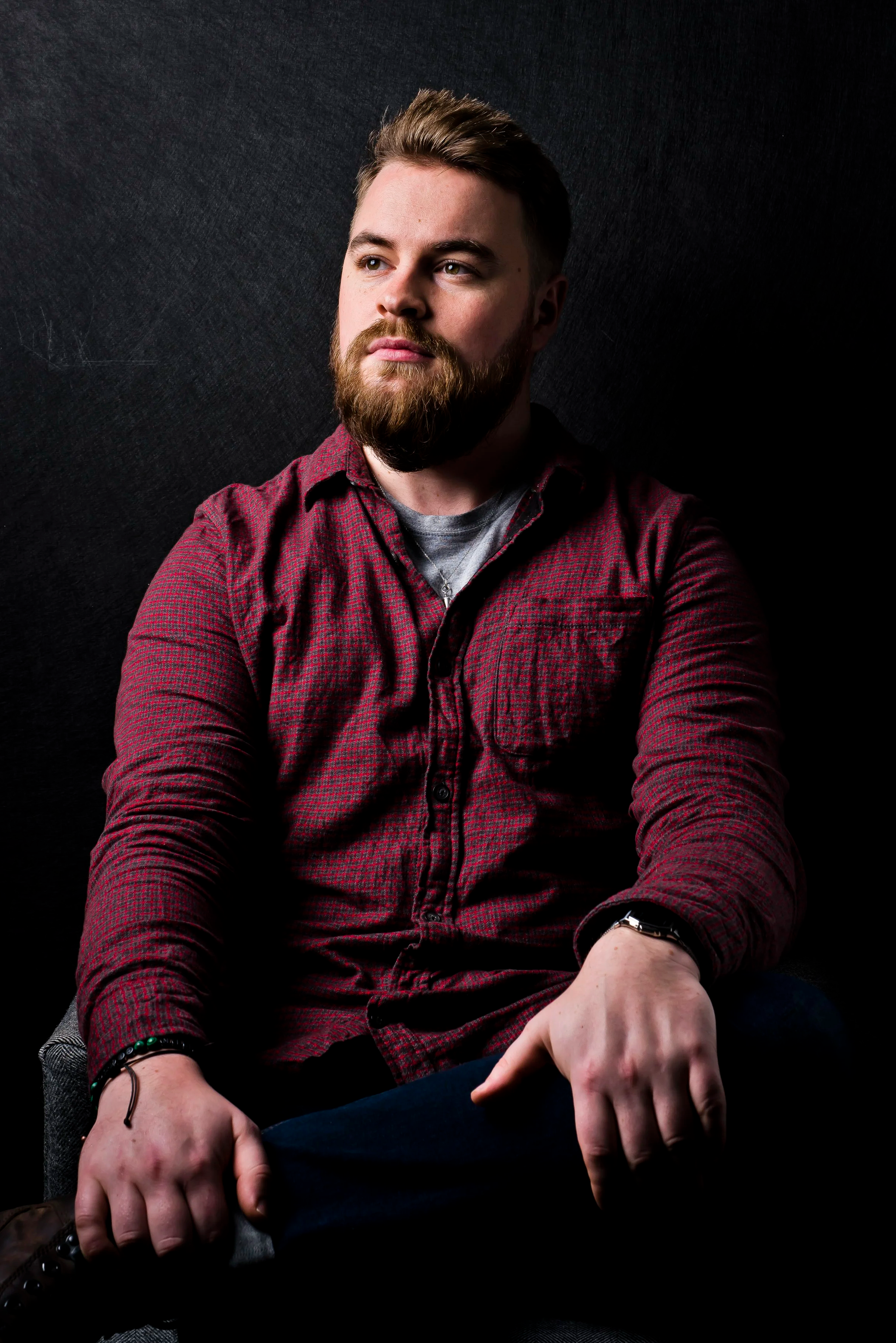 Portrait of a young man with light skin, a full beard, and styled brown hair, wearing a red plaid shirt over a gray t-shirt, with a watch on his left wrist, sitting against a dark background with muscular arms crossed over his lap.
