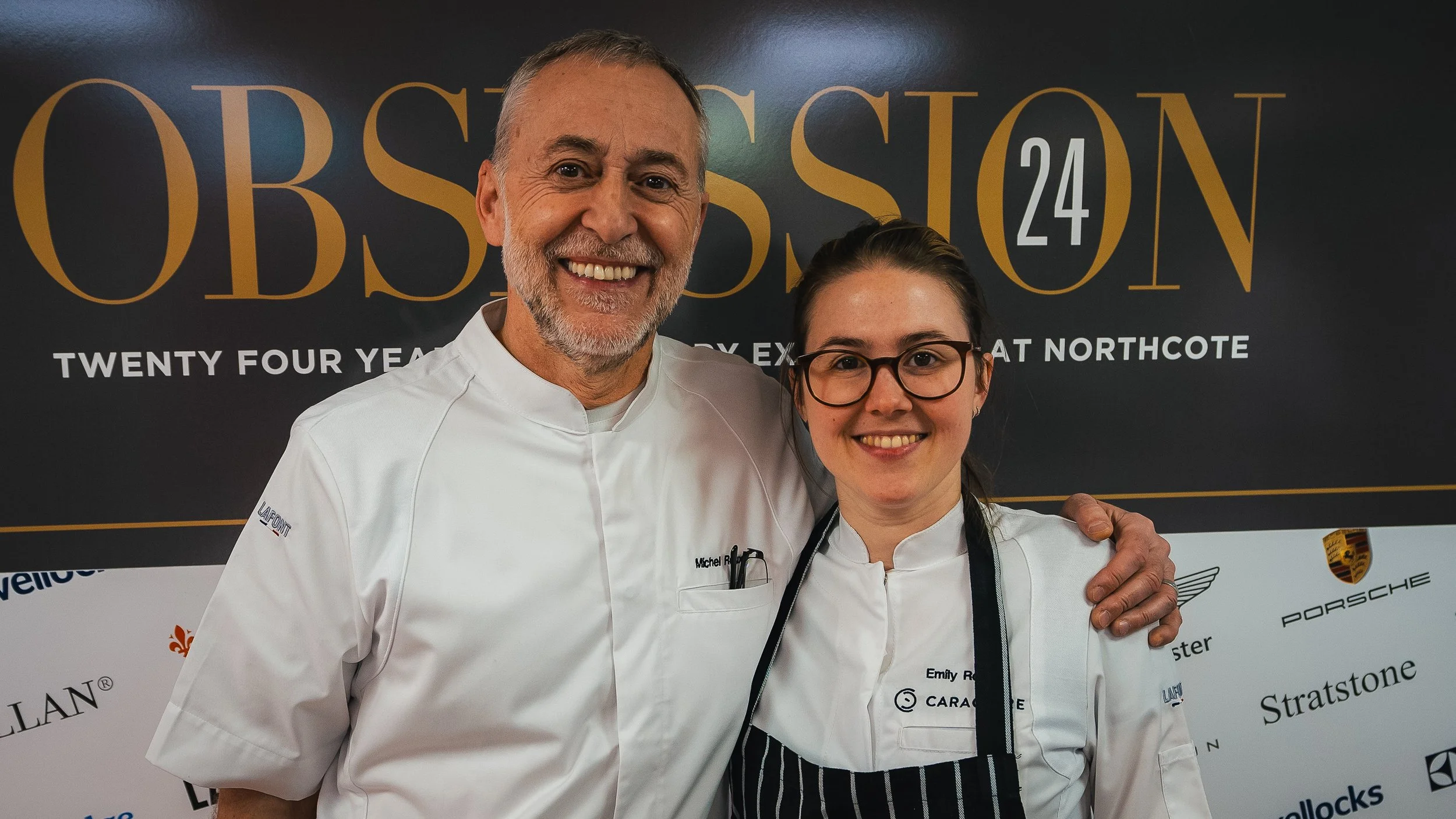 Two chefs in white uniforms smiling, standing in front of a black backdrop with gold and white text, at a professional event or competition.