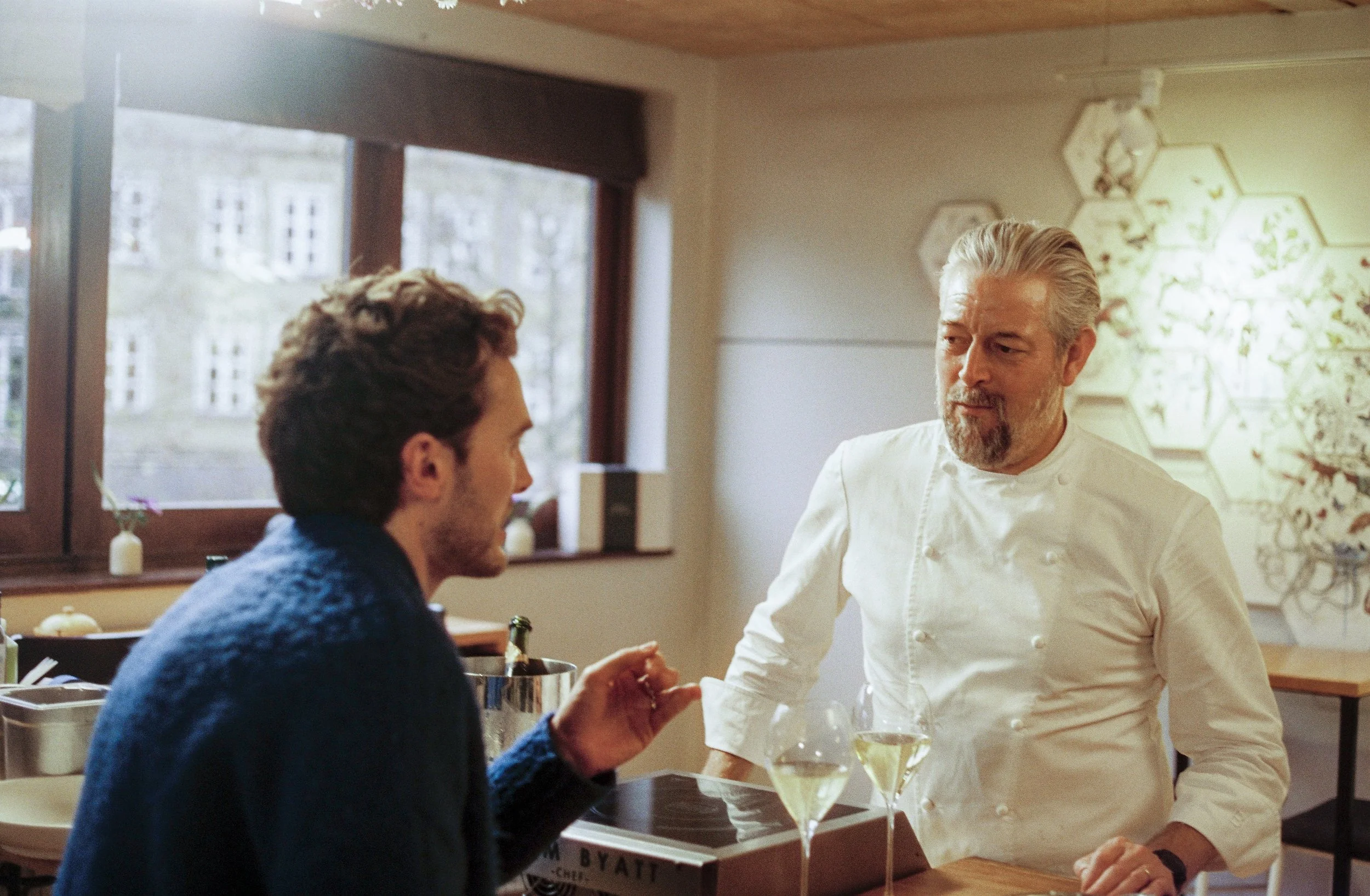 A chef in a white uniform and a man in a blue sweater having a conversation at a restaurant table with glasses of white wine. The setting is cozy with natural light, decorative wall art, and large windows.