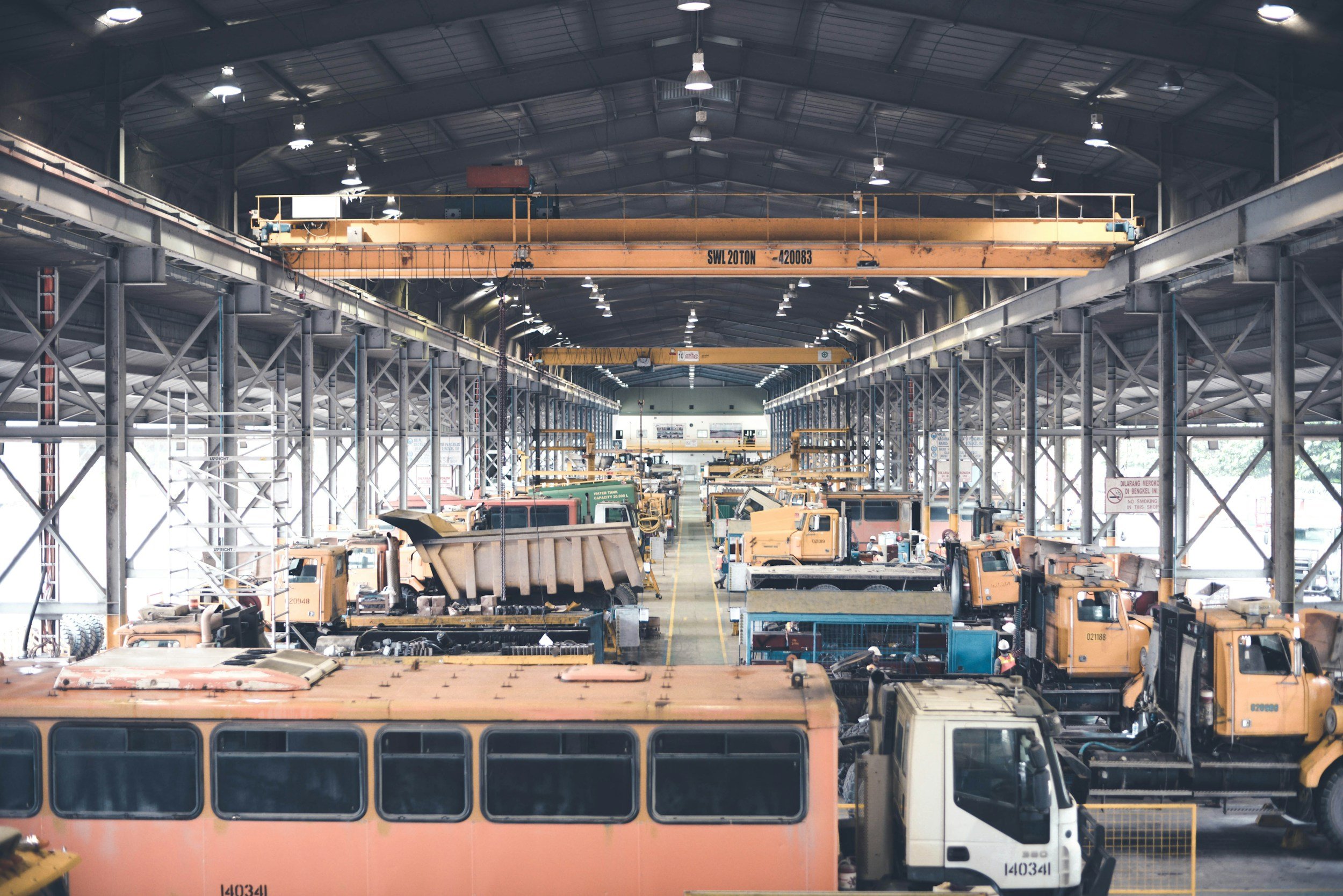 Interior of a large industrial warehouse with high metal roof, equipped with cranes and multiple construction vehicles.