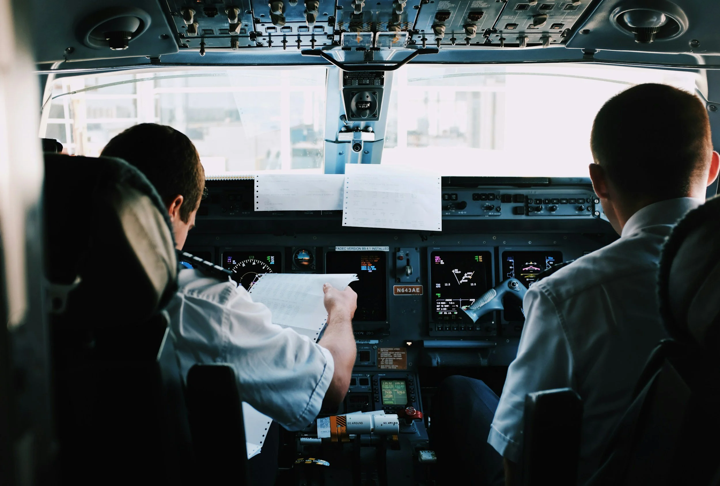 View from the cockpit of an airplane showing two pilots wearing white shirts, looking at flight instruments and checklists.