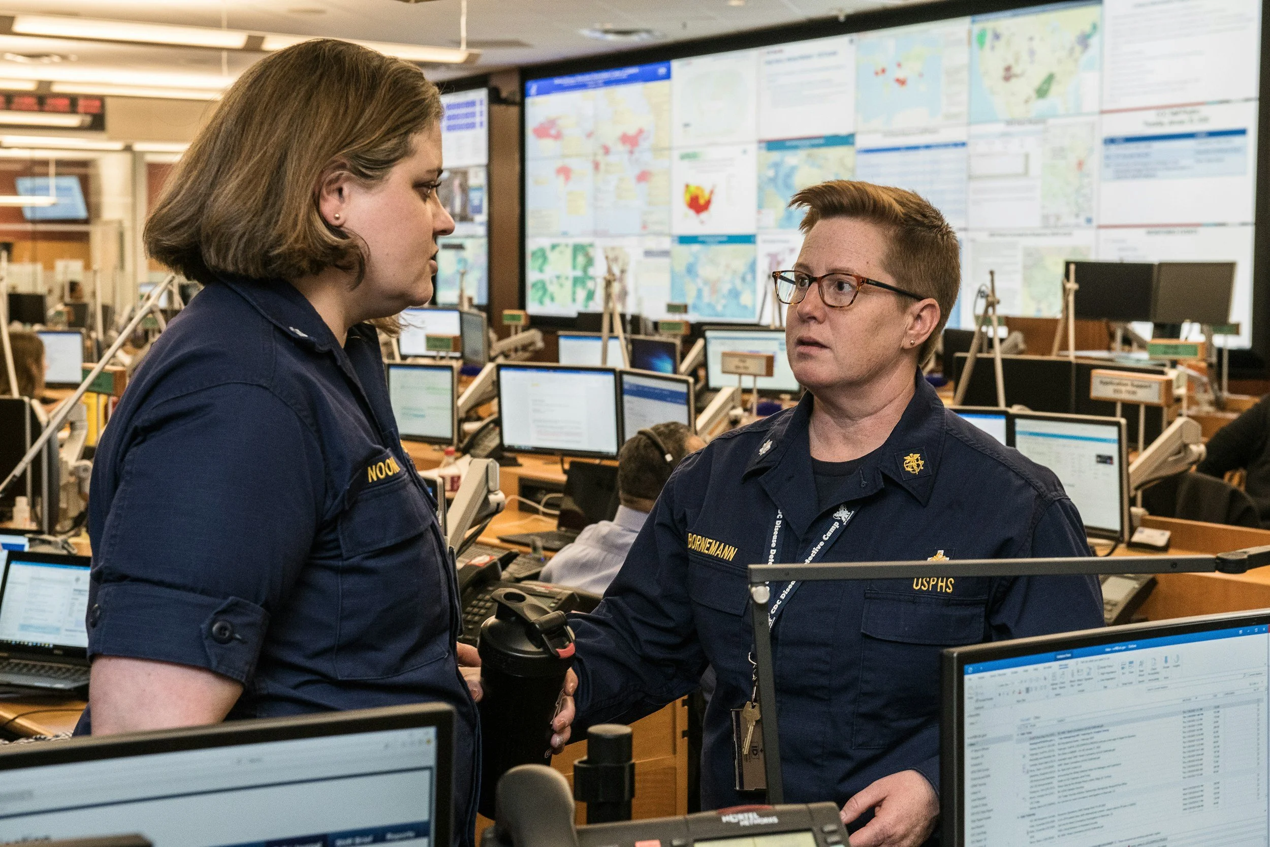 Two uniformed emergency response personnel, a woman and a man, having a serious discussion inside a busy operations center with multiple computer monitors, maps, and communication devices in the background.