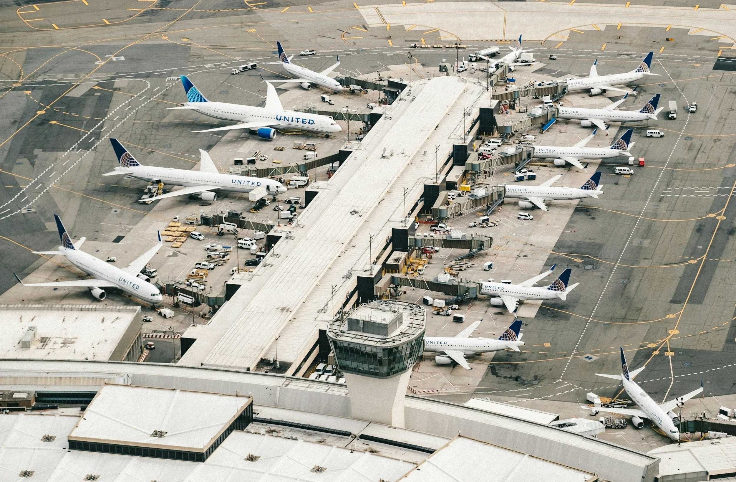 Aerial view of an airport terminal with multiple United Airlines airplanes parked at gates, connected by jet bridges, with ground service vehicles around.