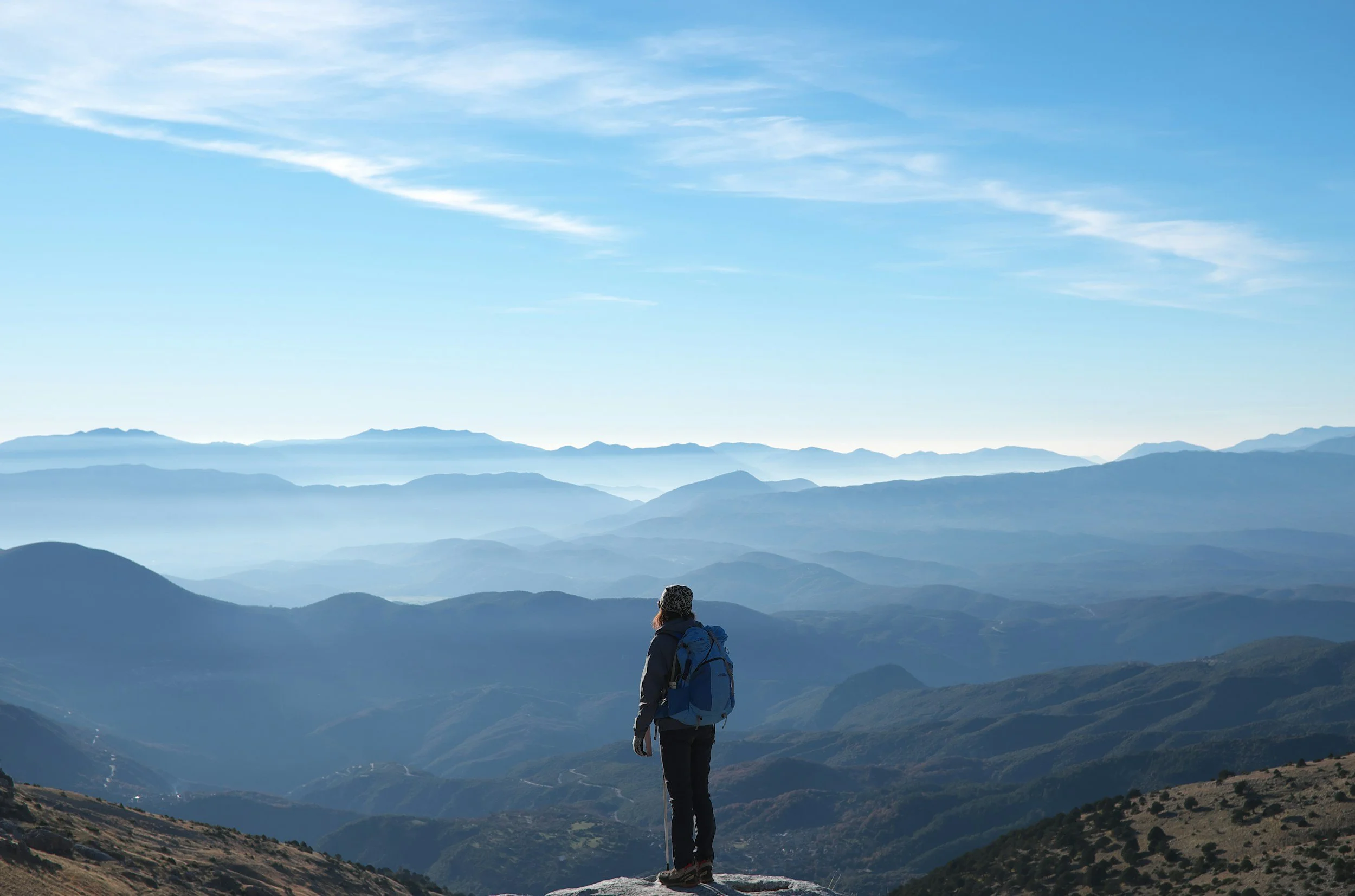 A person wearing a hiking backpack and hat stands on a mountain overlooking multiple mountain ranges, with a blue sky and wispy clouds above.