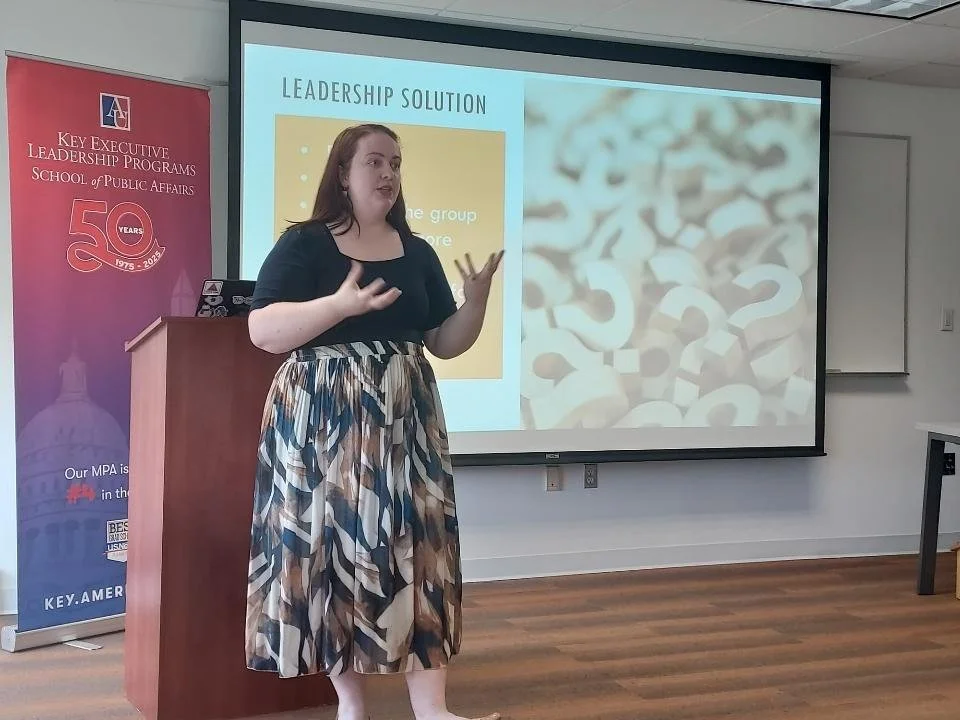 A woman giving a presentation in a classroom setting. She is standing next to a podium, gesturing with her hands. A large screen behind her displays a slide titled "Leadership Solution" with a blurred image of question marks and a bullet point list. To her left, there is a red banner celebrating 50 years of a leadership program, with the logo of a university.