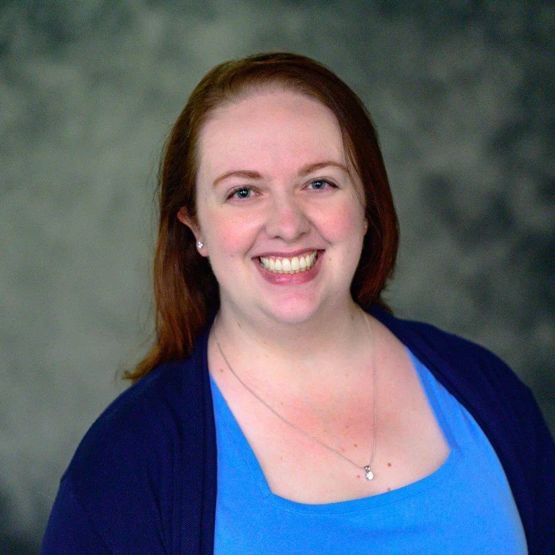 The Founder and Principal Claire Young, a woman with red hair is smiling, wearing a blue shirt and dark cardigan, against a gray mottled background.