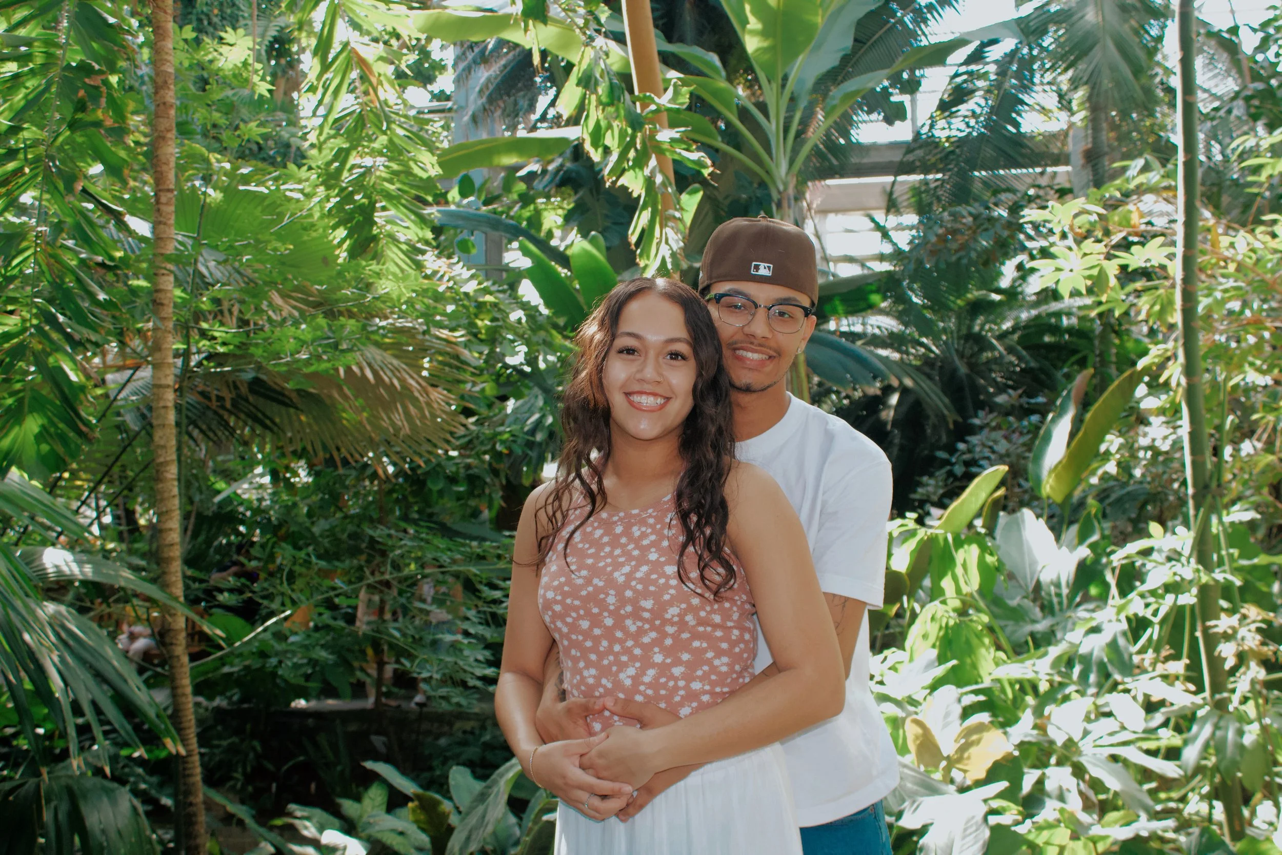 A smiling couple embracing in a lush indoor botanical garden with various green plants and trees.