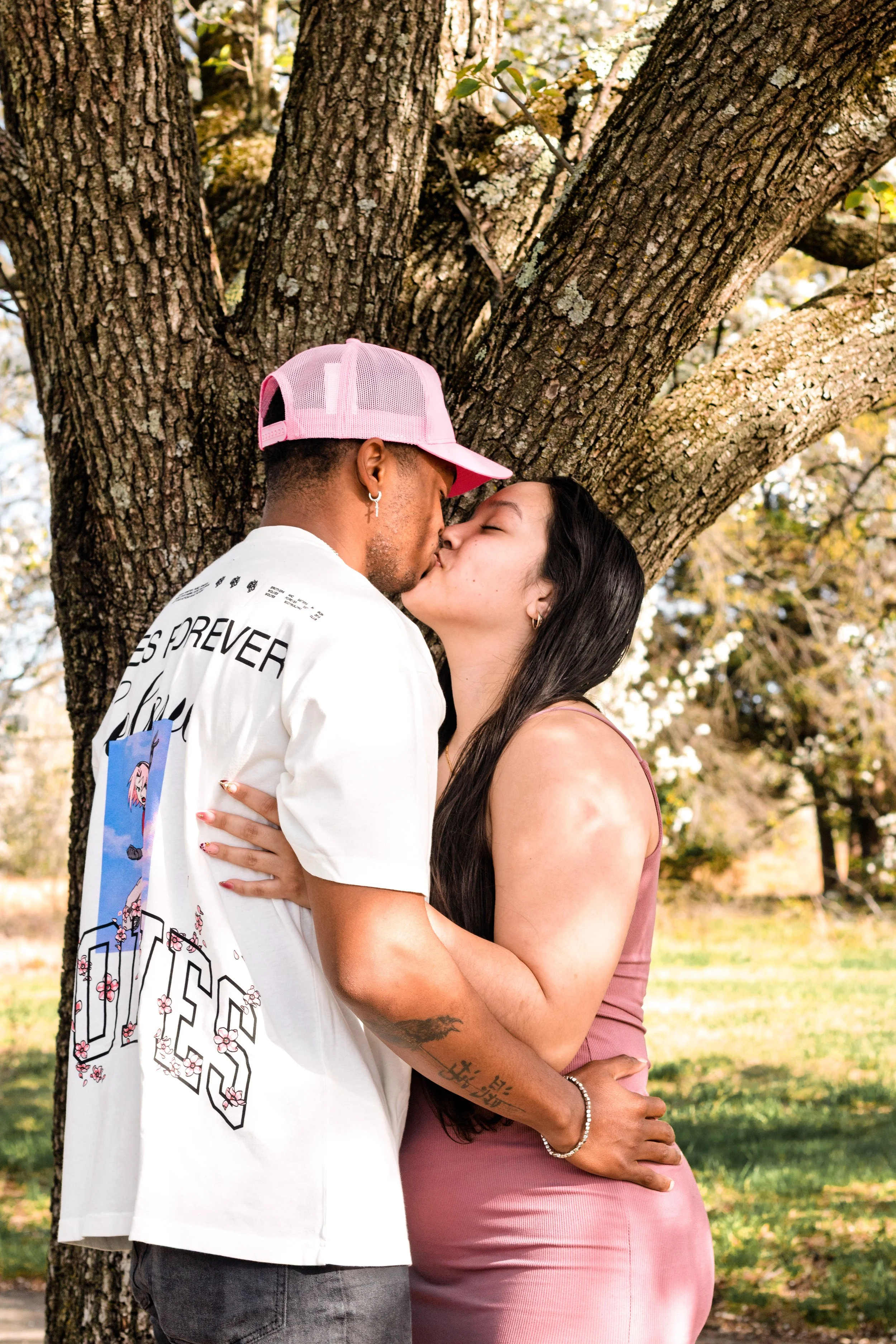 A couple sharing a kiss outside by a tree, with the man wearing a white shirt and pink cap, and the woman in a pink dress.