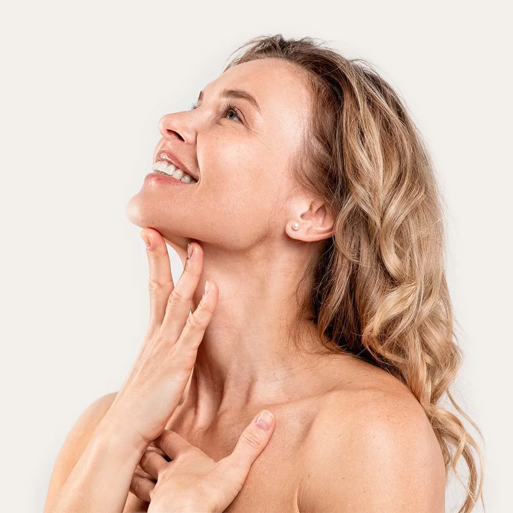 A woman with curly blonde hair smiling and touching her face and neck.
