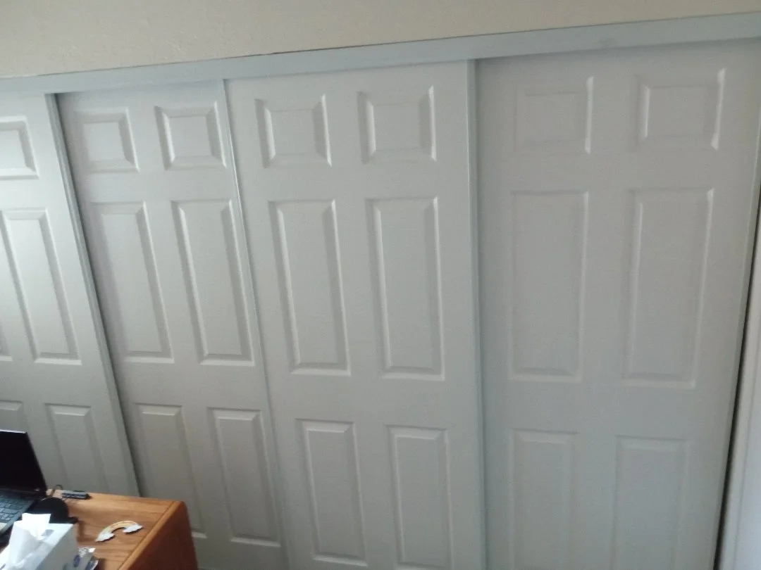 A wall with multiple white closet doors and a wooden desk with some items on it in the foreground.
