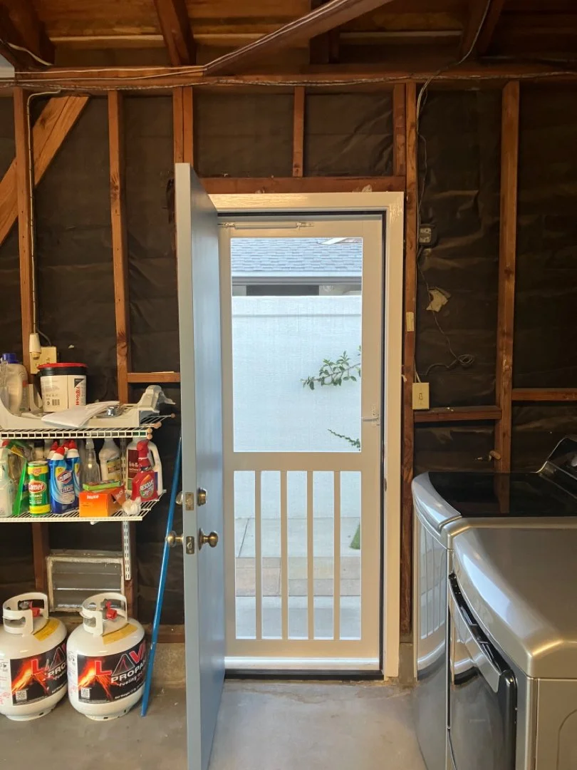 A garage with an open door leading outside, with laundry appliances, shelves with cleaning supplies, and propane tanks inside.