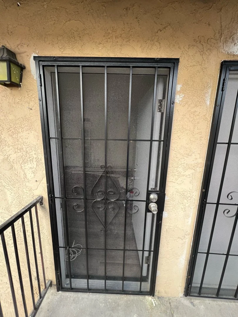 Outdoor black metal security door with a decorative heart-shaped design, leaded with a screen, attached to an exterior yellow stucco wall.