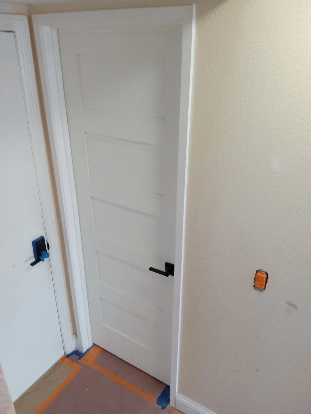 Interior of a room under construction with a white door, beige textured wall, electrical outlet box, and tape on the floor.
