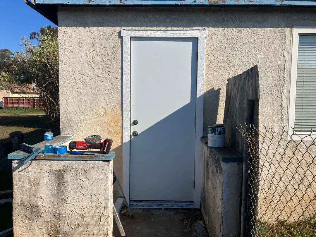 Backyard exterior with a white door on a beige textured wall, tools and paint supplies on a concrete surface, a chain-link fence, and a window with blinds.