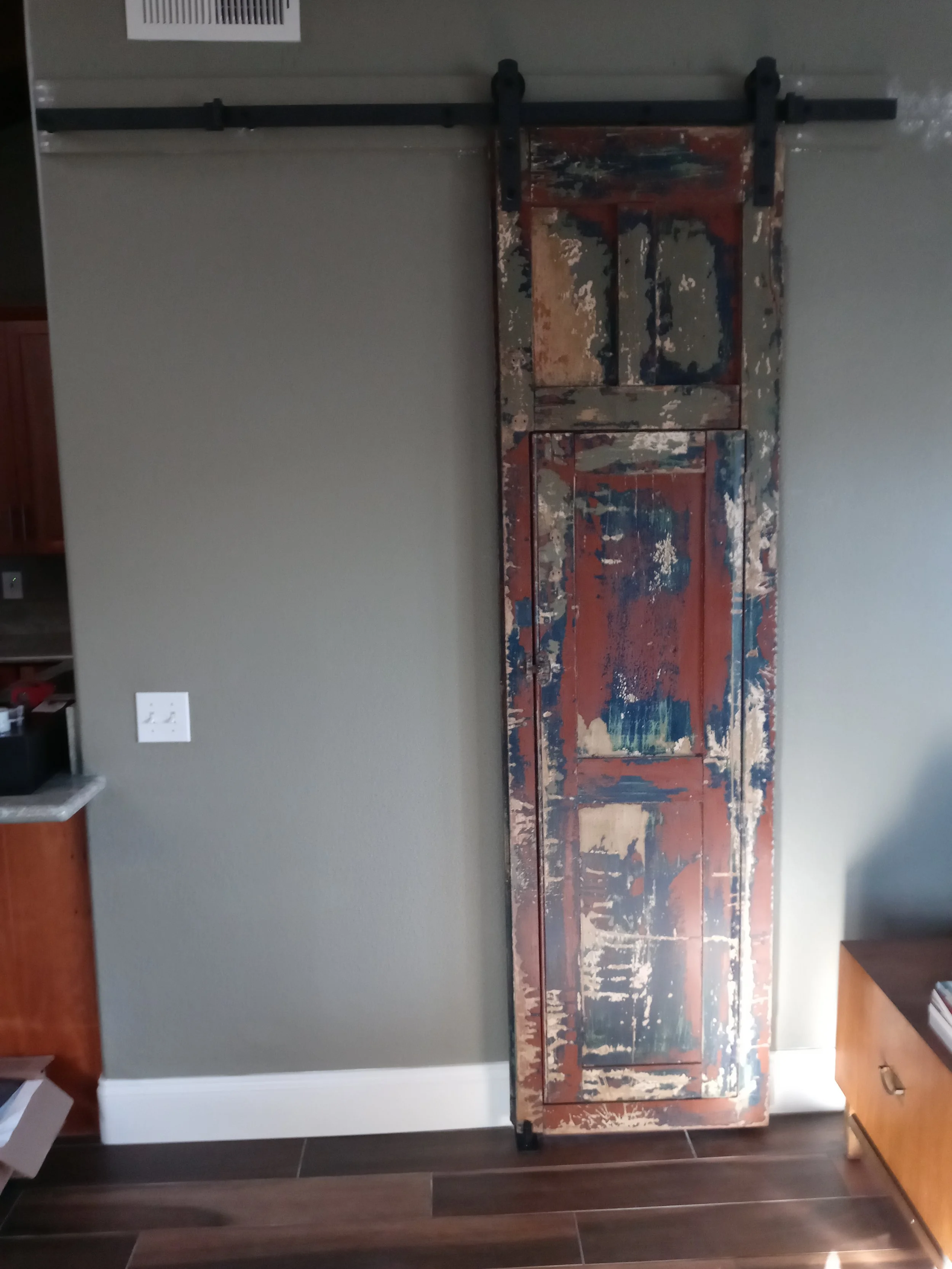 A rustic, weathered wooden barn door with chipping paint in various shades of red, blue, and beige, hanging on a black sliding track on a gray wall.