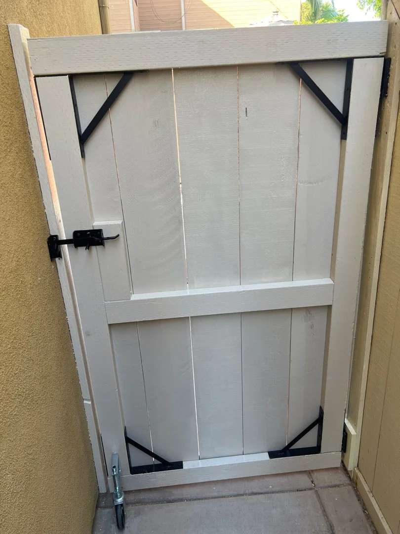 A small outdoor gate made of light-colored wooden planks reinforced with black metal brackets, secured with a latch, and mounted between two walls.