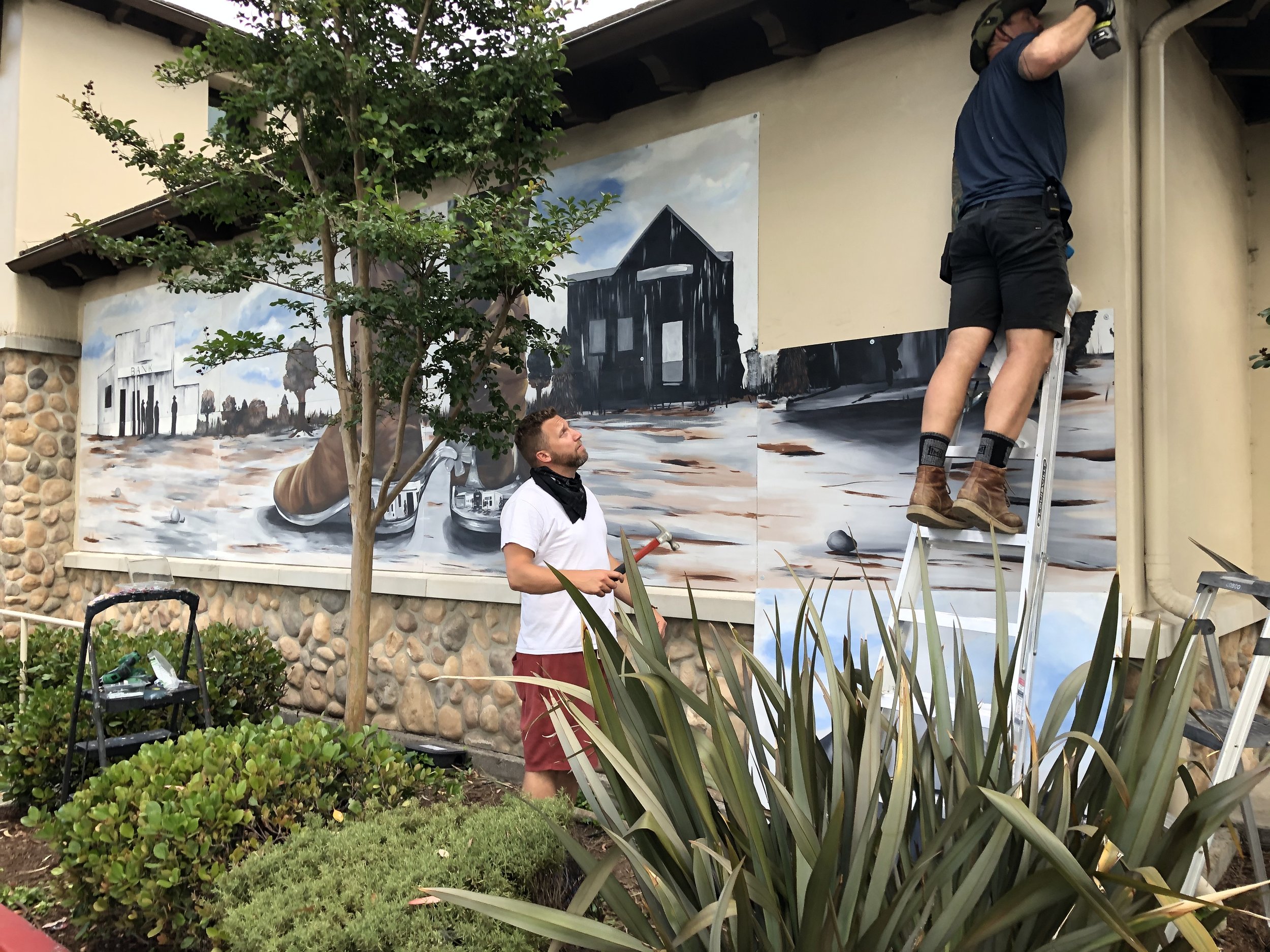 Two artists working on a large mural outdoors. One artist is standing on a ladder, painting a black building, while another artist watches. The mural depicts a scene with a large boot and a building in a somewhat abstract style. There are bushes, a tree, and gardening tools around them.