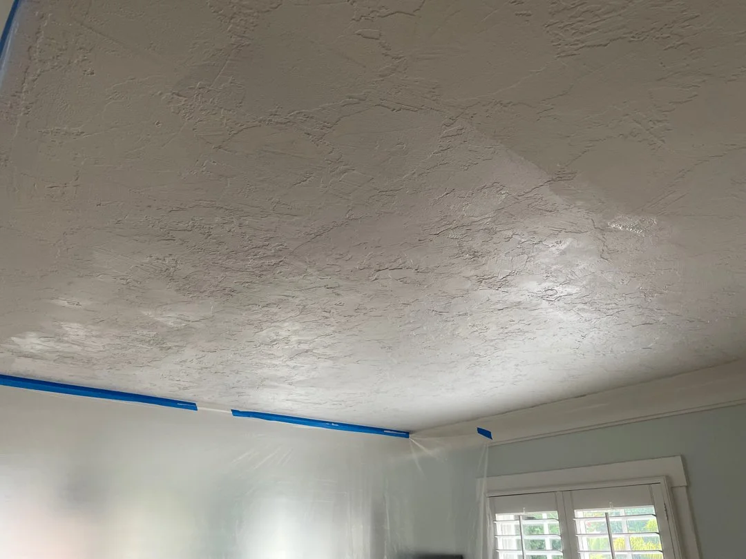 Ceiling with textured finish in a room under renovation, blue painter's tape along the edges, window with blinds in the background.