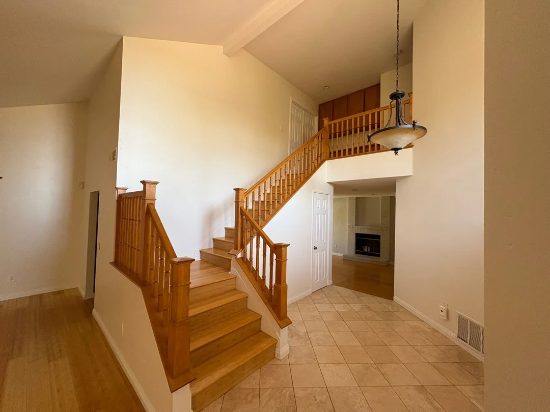 Interior of a house showing a staircase with wooden banisters, a hanging light fixture, a white door, and a corner with a fireplace in a room.