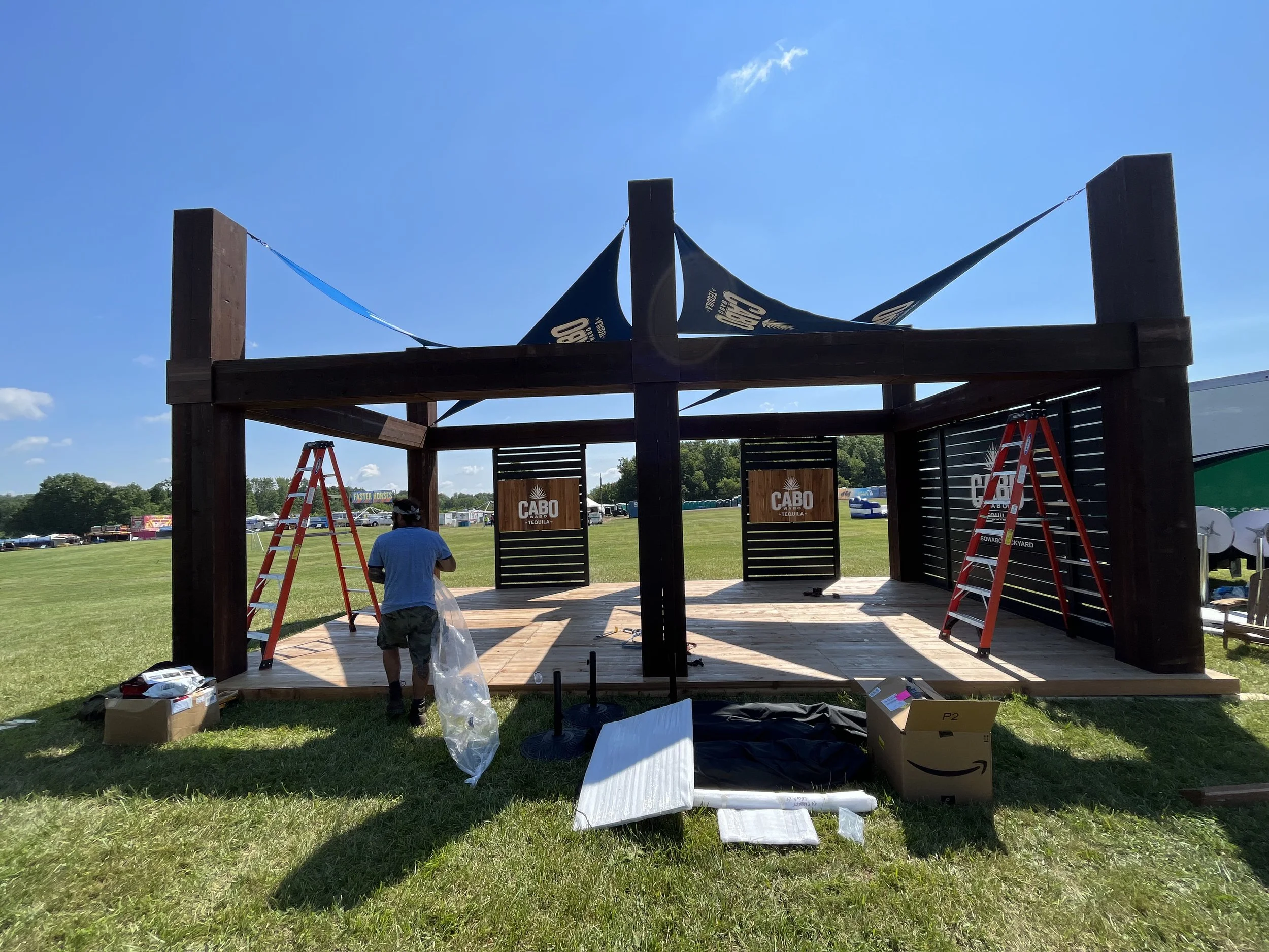 A partially assembled outdoor stage structure with a wooden floor, supported by large wooden posts, set up on a grassy field under a clear blue sky. There are ladders, boxes, and tools around, with black and wooden panels displaying the logo 'Cabo Tequila' on the stage backdrop. A man is seen working on the stage.