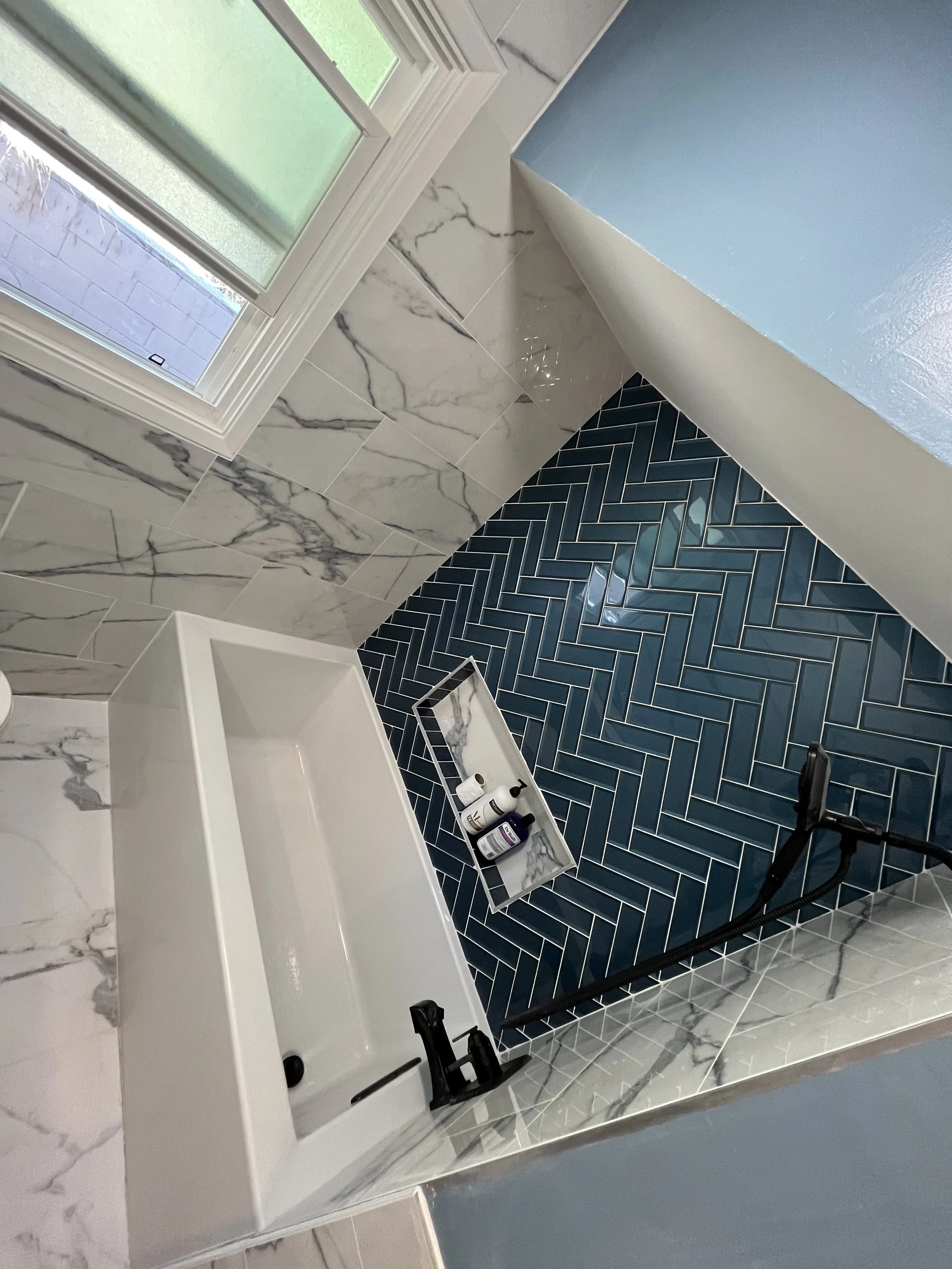 Bathroom with blue herringbone tile floor, white vanity with marble countertop, marble wall tiles, and a small shelf holding bottles. A window with frosted glass is visible.