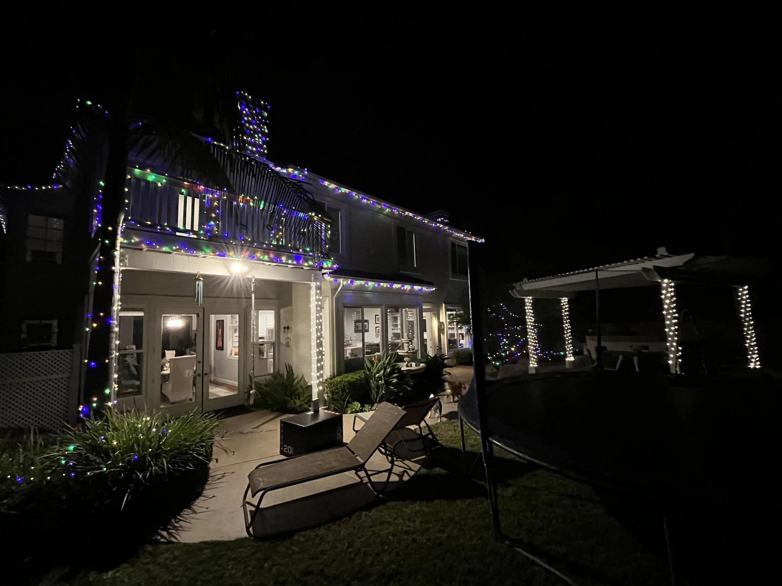 House decorated with colorful string lights at night, with patio furniture, plants, and a trampoline in the yard.