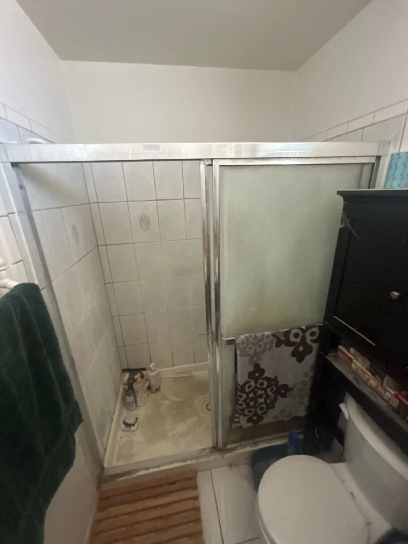 A small bathroom shower area with a corner shower stall, frosted glass door, and beige tile walls. Items such as shampoo bottles are on the floor inside the shower. To the right, part of a toilet with a cabinet above it is visible, and a towel hangs on the wall to the left.