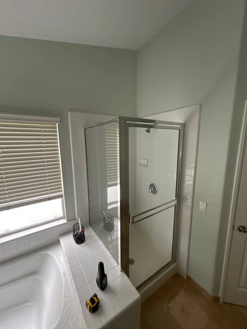 Bathroom with large window covered by blinds, corner shower with glass door, white tile surfaces, and a few tools placed on the edge of the bathtub.