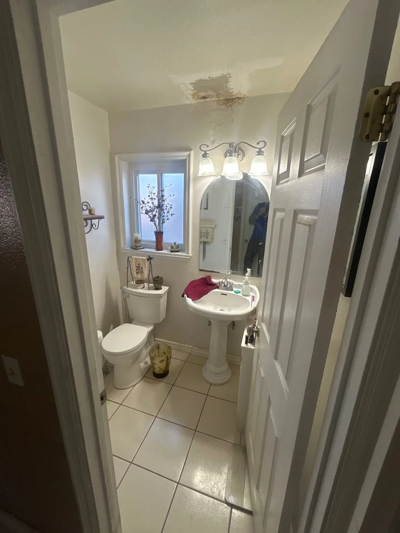 Small bathroom with white walls and tile floor. There is a window with a small plant, a toilet, a pedestal sink with a mirror and three light fixtures, and a small trash can. The ceiling has a water stain.